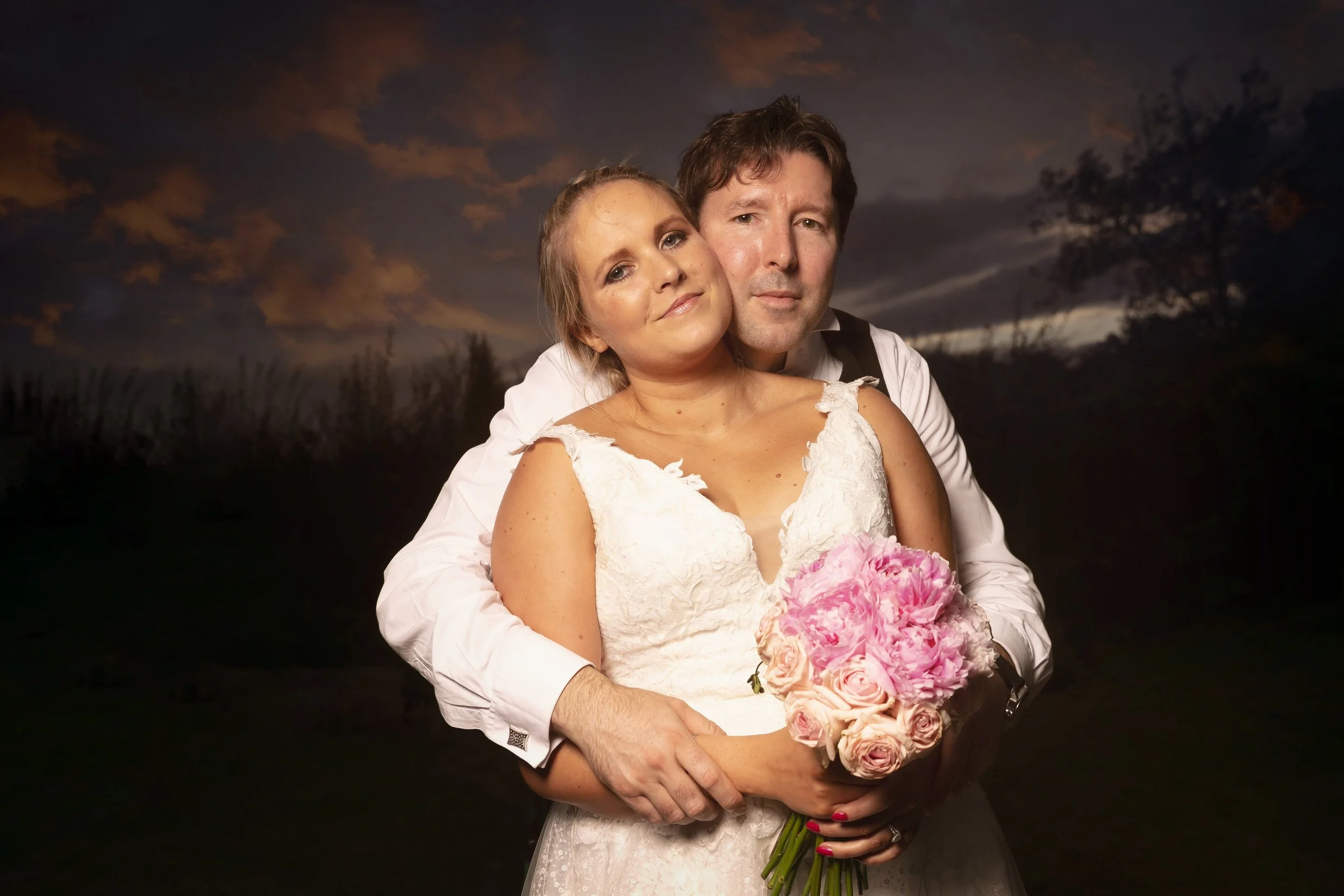 A couple on their wedding day stands together outdoors at sunset, the woman in a white wedding dress holding a pink and white bouquet, the man in a white shirt and black vest embracing her.