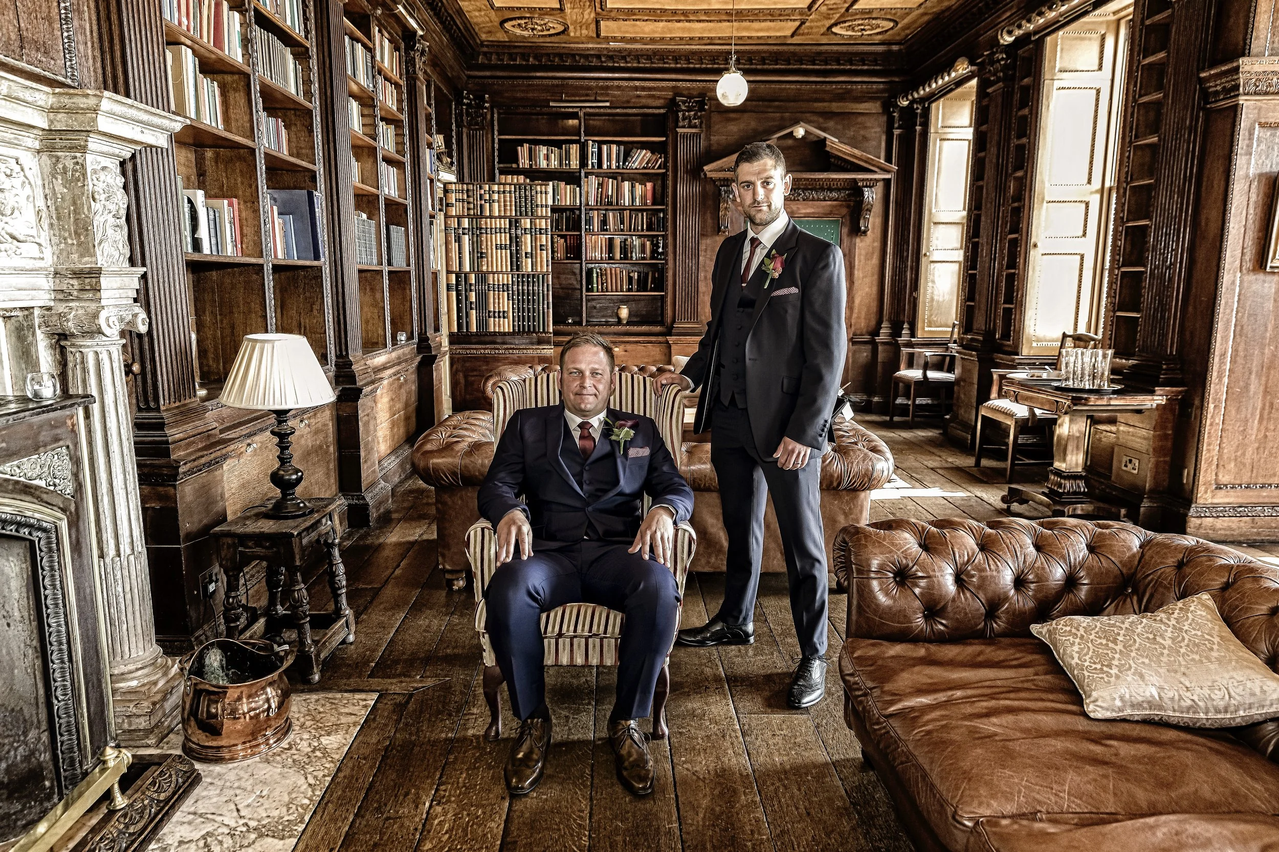 Two men dressed in suits, one seated in a striped armchair and the other standing with a hand on the seated man's shoulder, are inside a richly decorated wood-paneled room filled with books and vintage furniture.