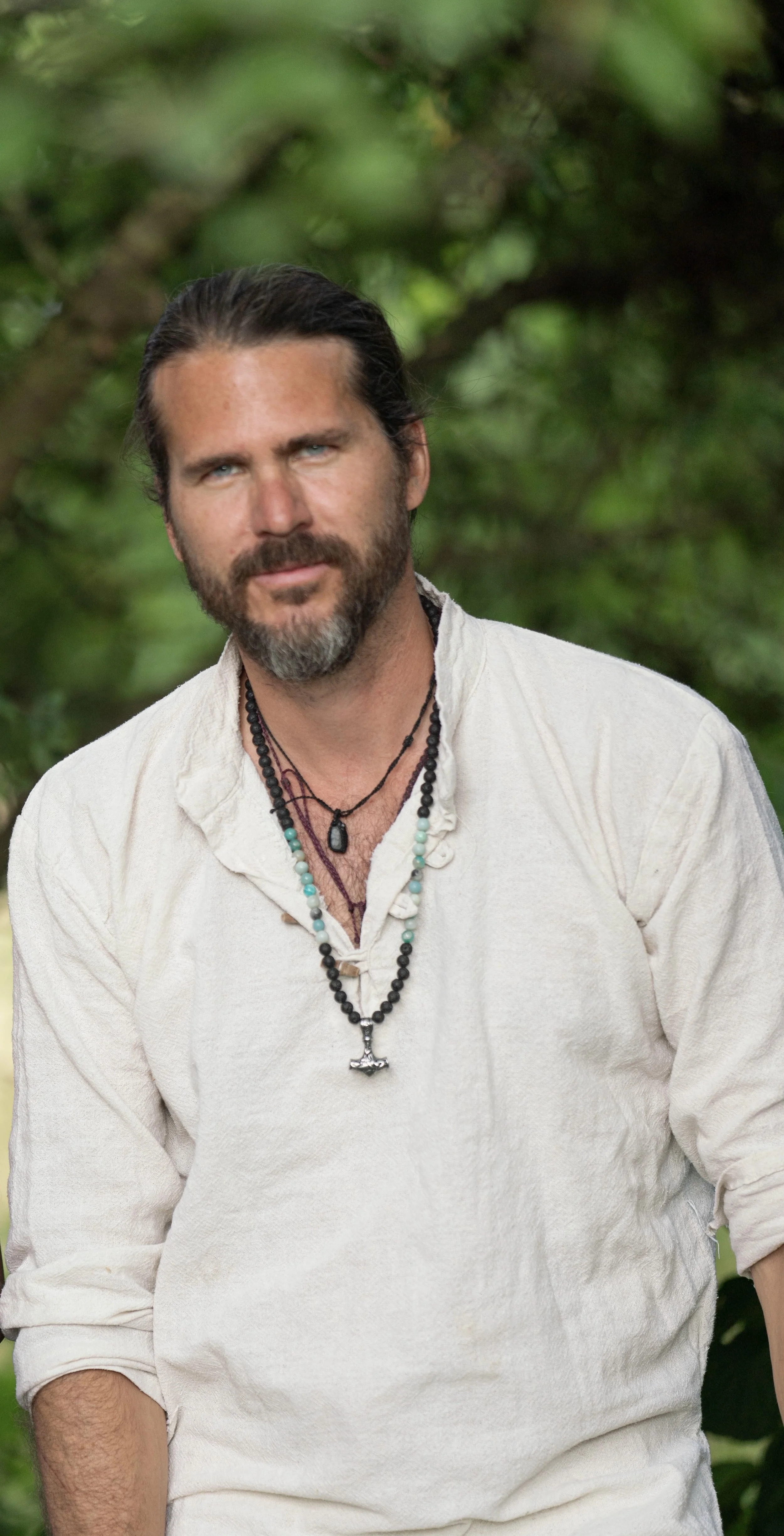 Ein Mann mit dunklem Haar, Bart und blauen Augen trägt ein weißes, lockeres Hemd und mehrere Halsketten, steht im Grünen