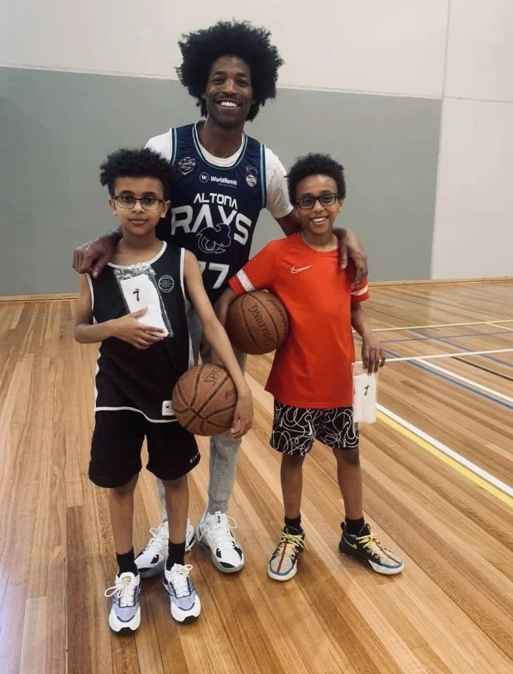 A man and two boys standing on a basketball court. The man is smiling, has afro hair, and is wearing a blue Altona RAYS jersey, with his arm around the boys. Both boys are holding basketballs and certificates, wearing athletic clothes and sneakers. The boy on the left has glasses and a black jersey, and the boy on the right is in an orange shirt and patterned shorts.