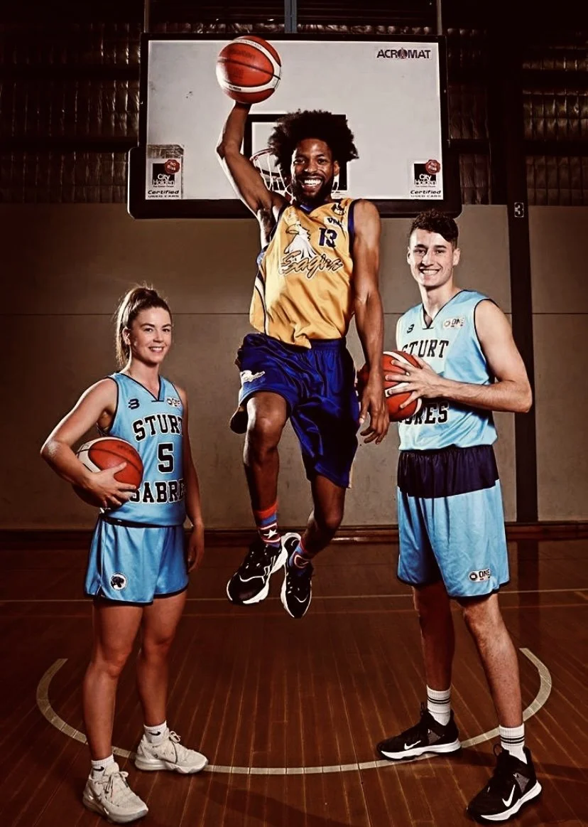 Three basketball players on an indoor court in front of a hoop, two in blue jerseys and one in a yellow jersey. The player in yellow is jumping and holding a basketball above his head. The two players in blue are holding basketballs and smiling.