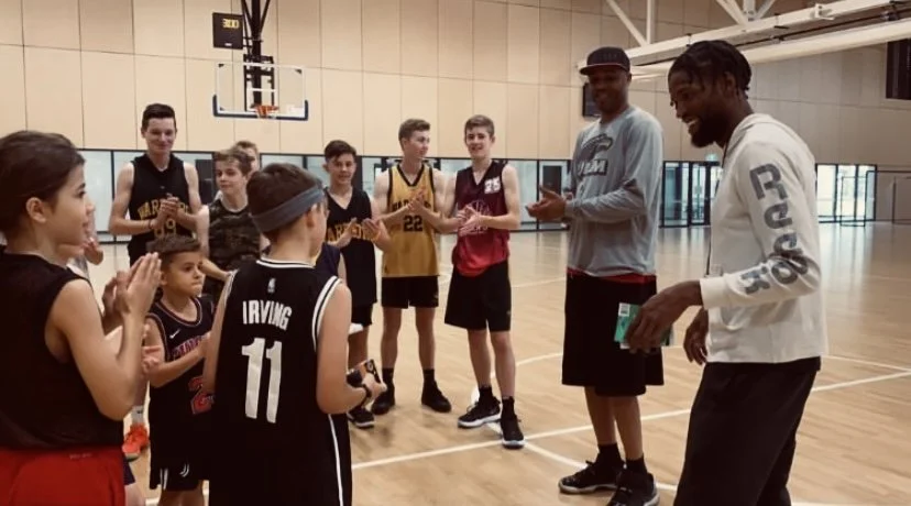 A group of young basketball players, including children and teenagers, gathered in a gymnasium around two tall men who appear to be coaches or trainers, engaging in conversation.