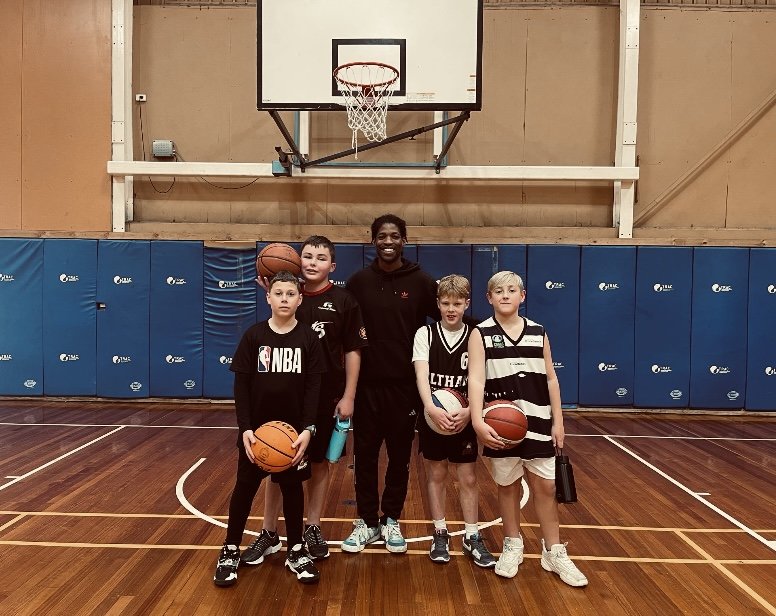 Five boys and one adult standing on a basketball court with basketballs in their hands, smiling, in front of a basketball hoop.