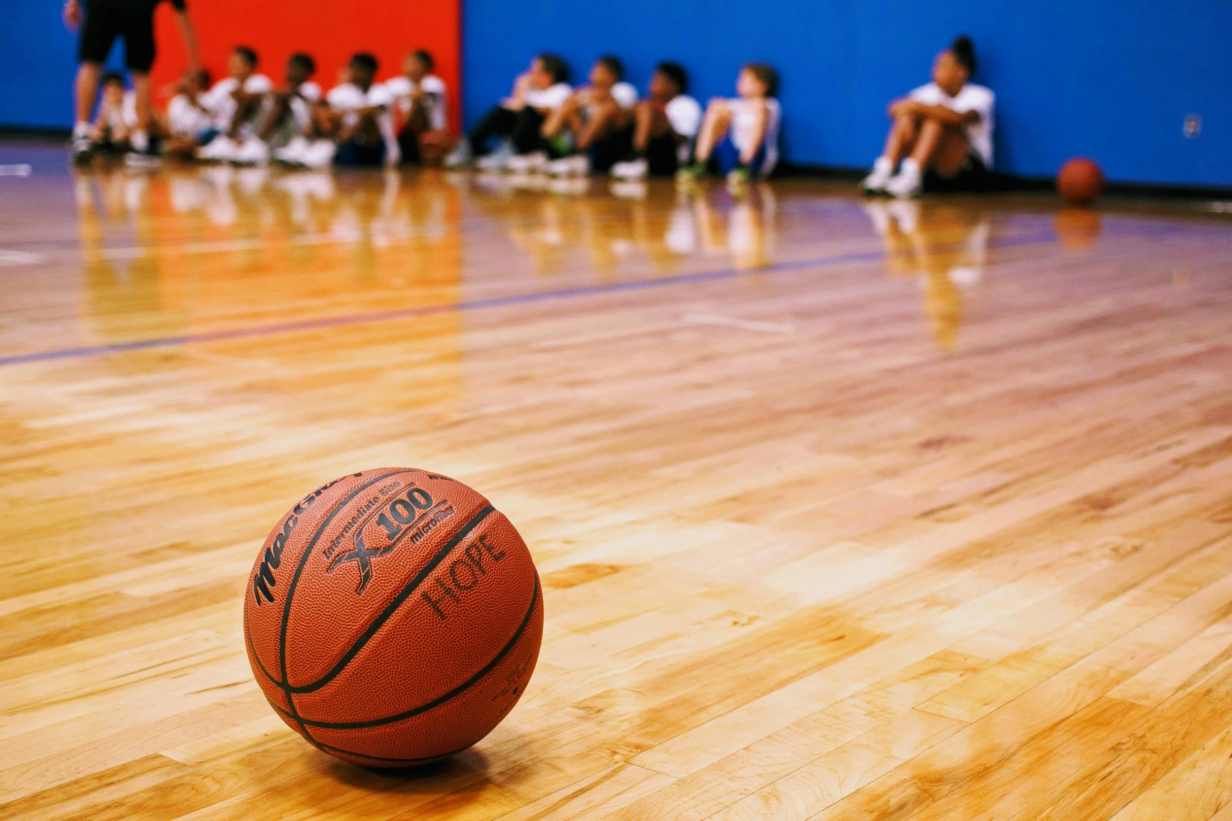 An orange basketball on a wooden gym floor with children sitting against a blue wall in the background.