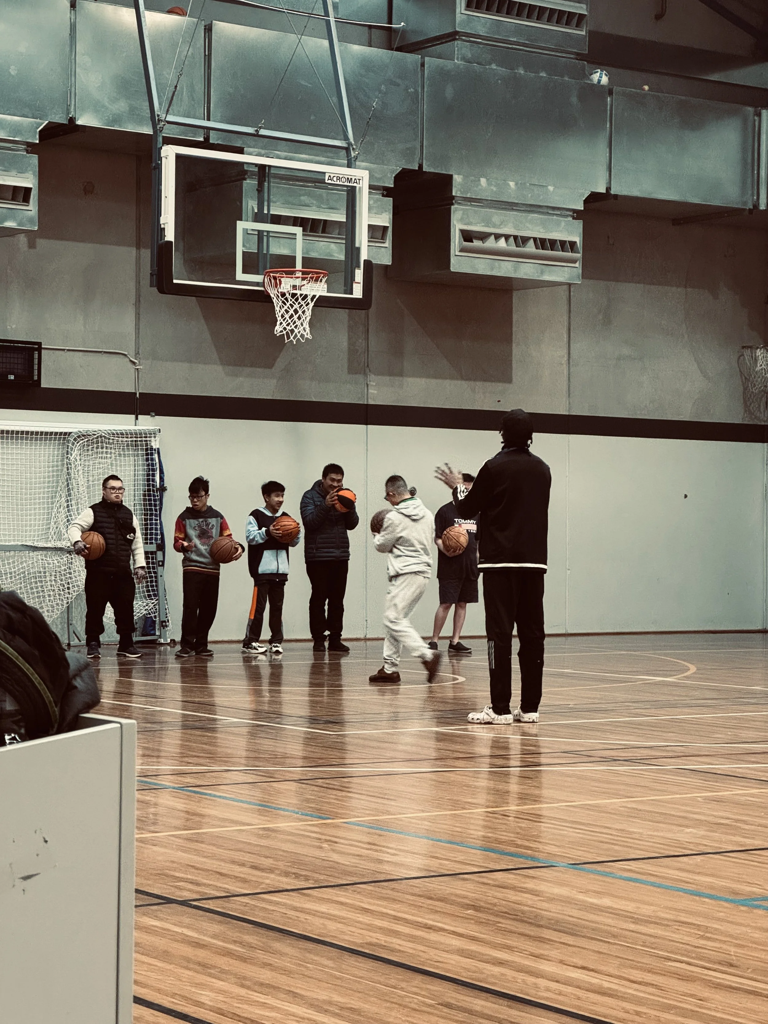 A basketball coach instructs a group of young children during a practice session in an indoor gymnasium. The children are holding basketballs and are standing in a line facing the coach, who is explaining a drill or technique.
