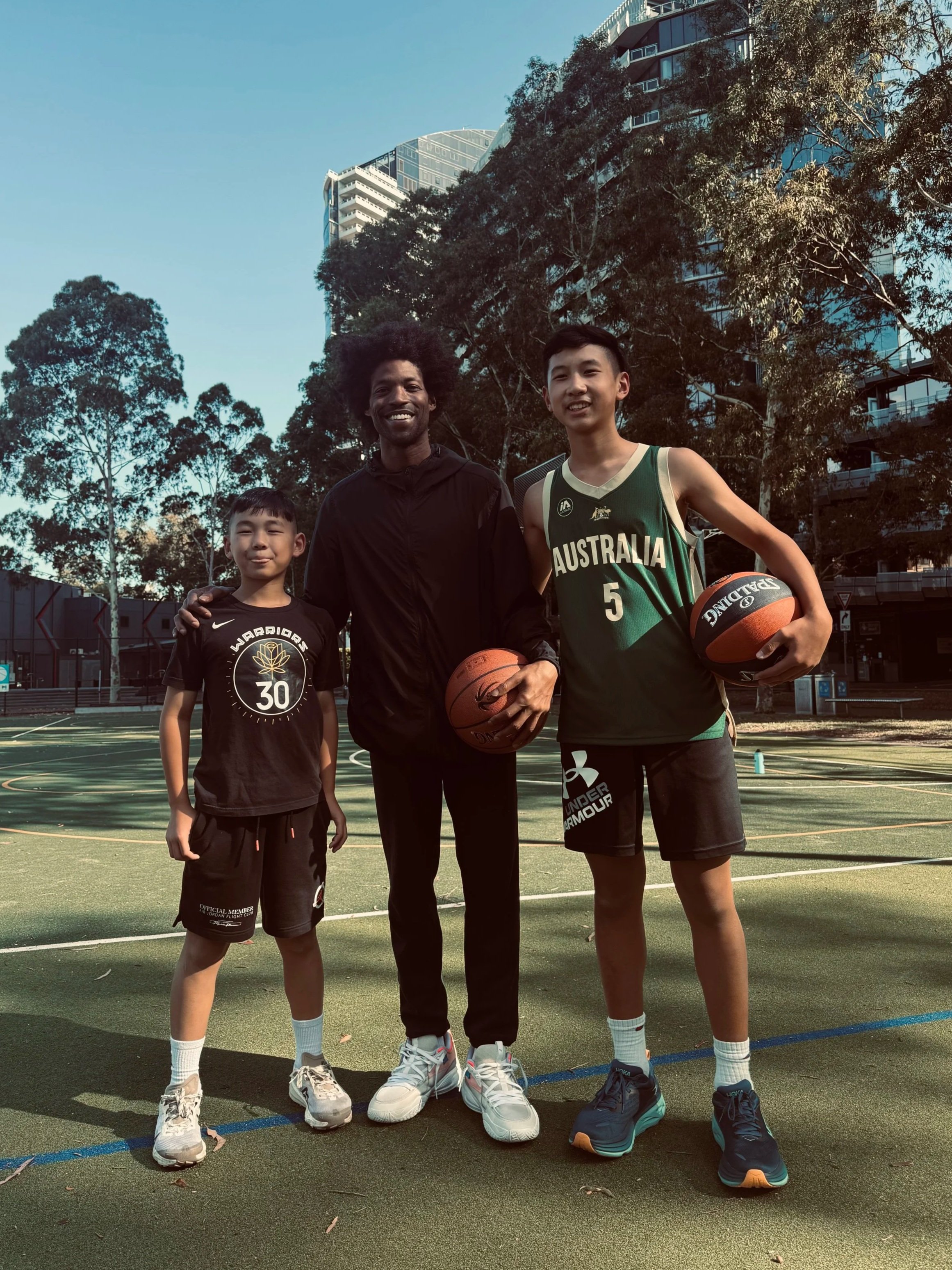 Three males standing on an outdoor basketball court, holding basketballs, smiling, with trees and high-rise buildings in the background.