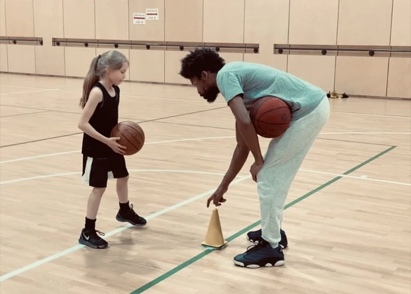 A young girl and an adult man practicing basketball drills in an indoor gym. The girl is holding a basketball, and the man is adjusting a cone on the floor as part of a training exercise.