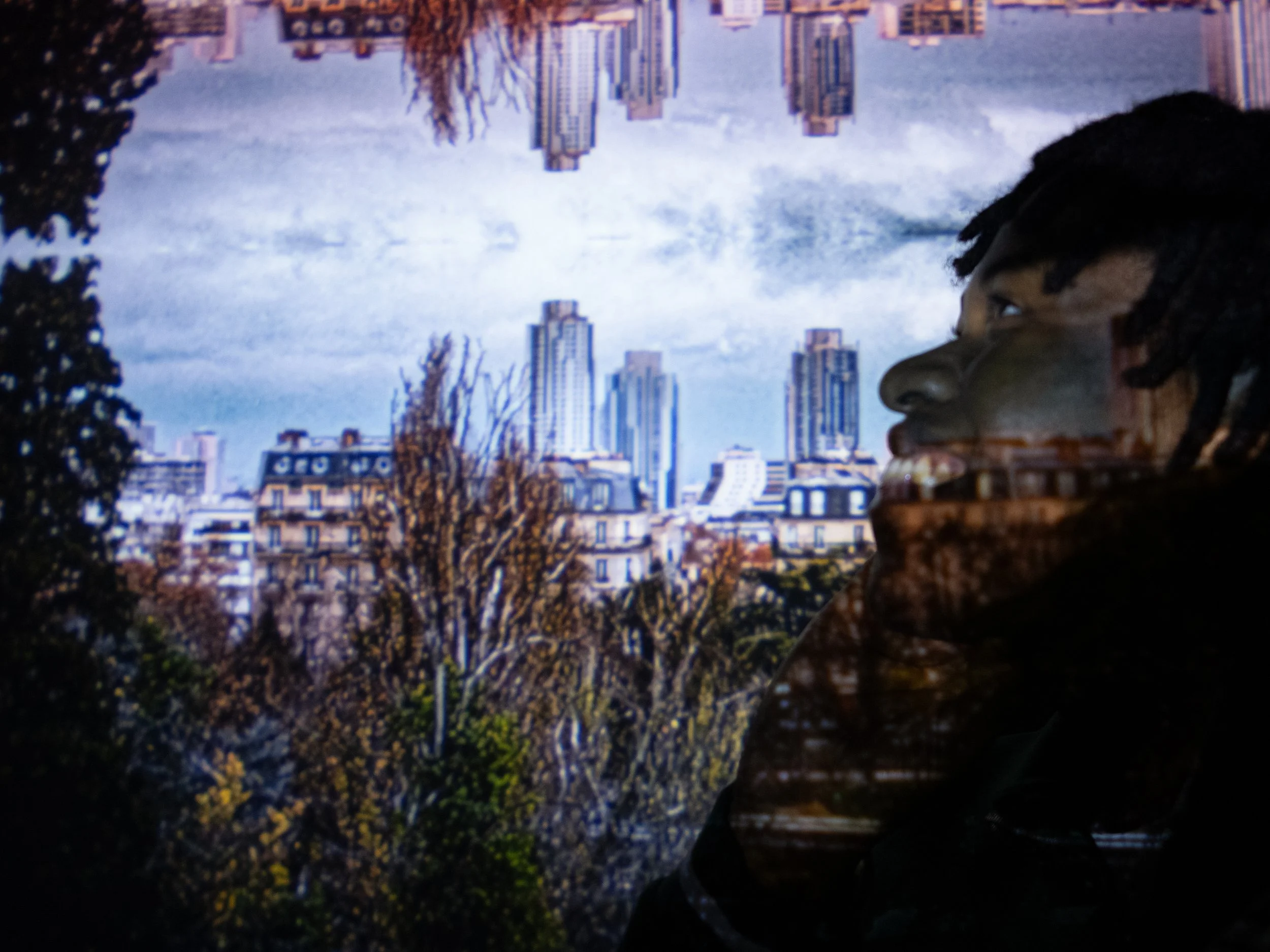 Photographe portrait d'intérieur sous base lumière photo artistique en Essonne Un portrait d'une personne avec des dreadlocks, avec la ville en arrière-plan, avec des bâtiments modernes et des arbres, vue à travers une surface réfléchissante.