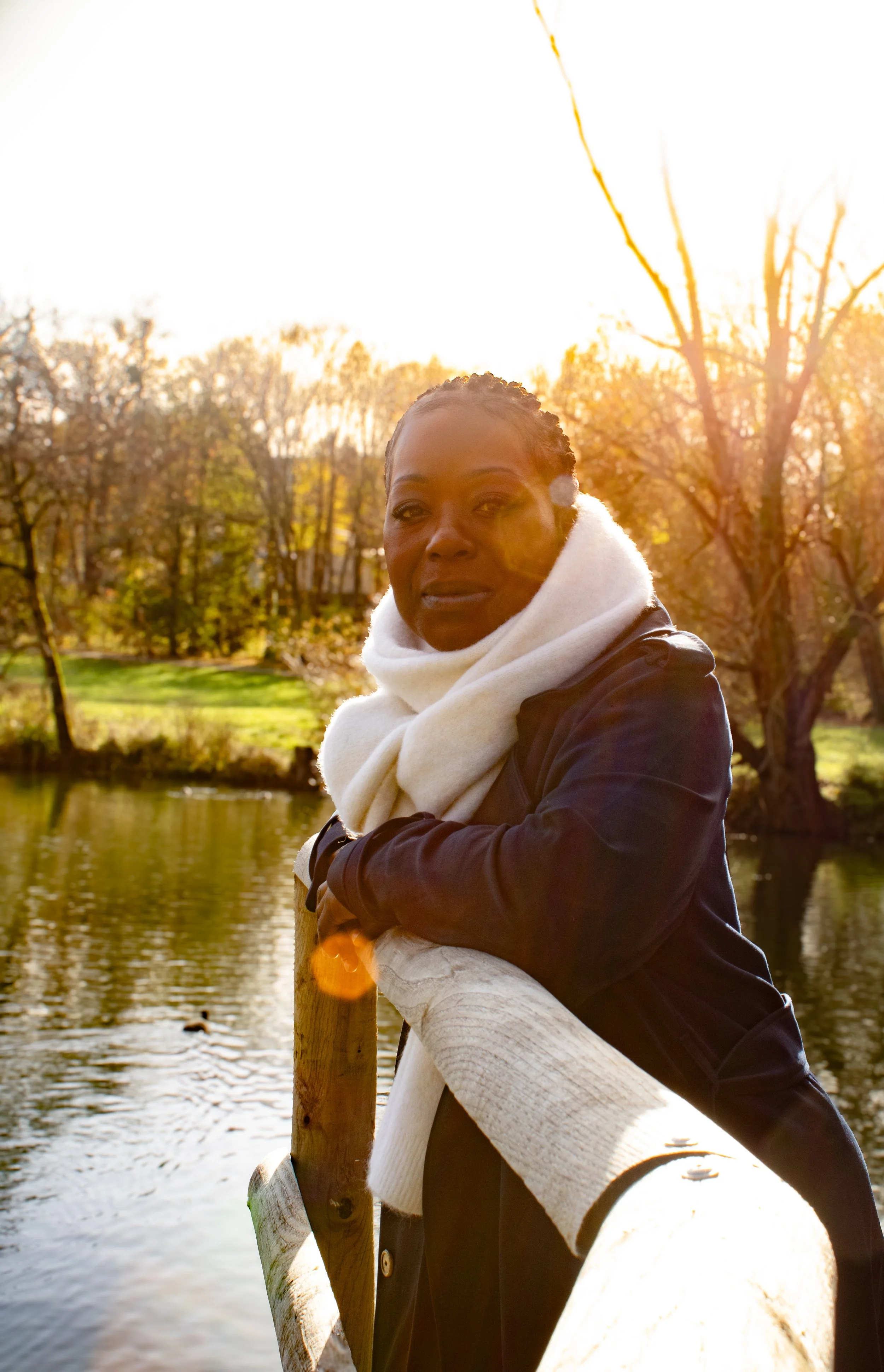 portrait femme dehors Essonne 91 photographe famille  Femme regardant la caméra, debout sur un pont en bois au bord de l'eau, en automne, avec des arbres aux feuilles jaunes et marron, soleil levant ou couchant derrière elle.
