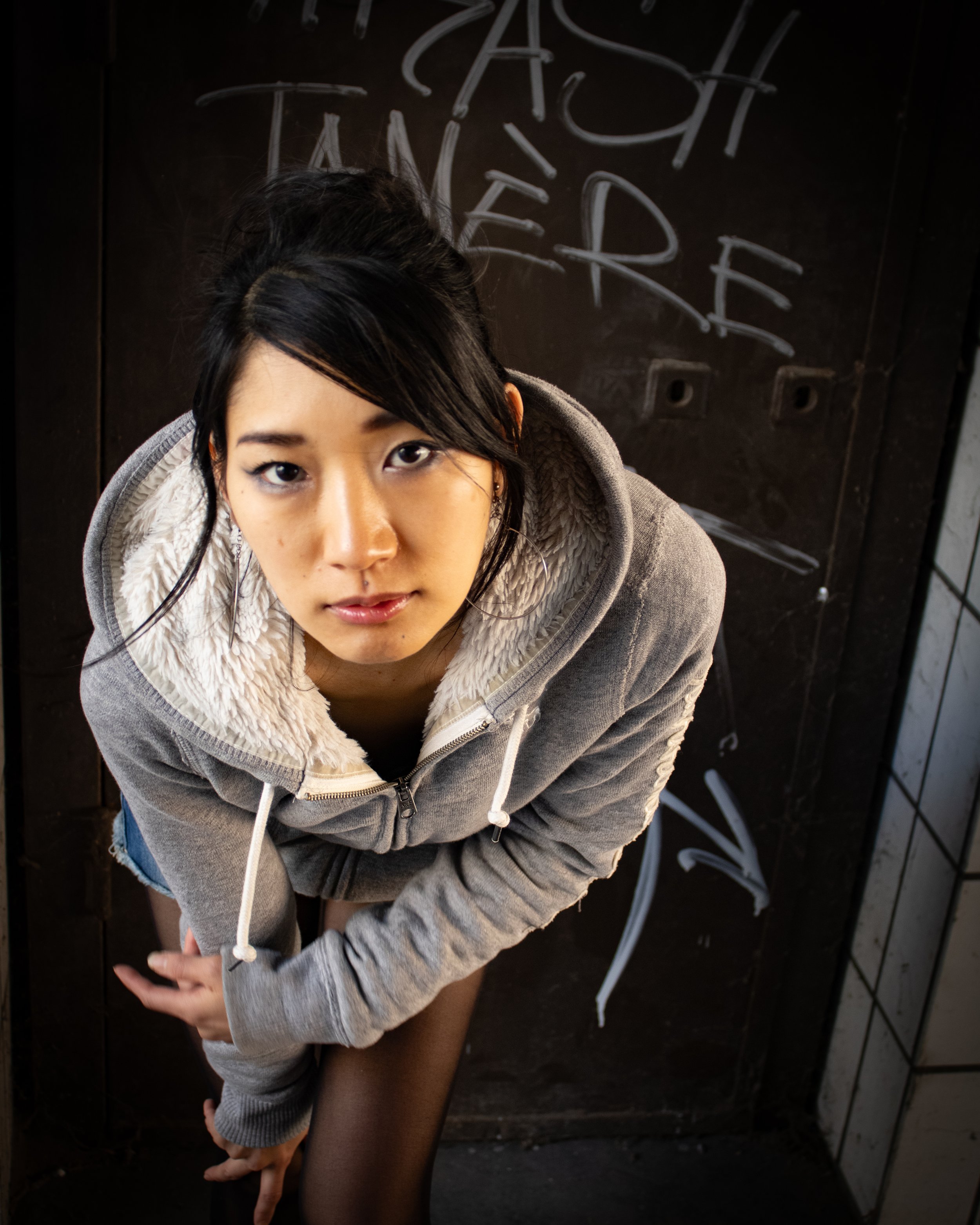 photo streetwear d'un photographe de portrait  Jeune femme regardant vers l'appareil, prise de vue en plongée, portant un sweat à capuche gris à l'intérieur d'une pièce avec un mur noir derrière elle.