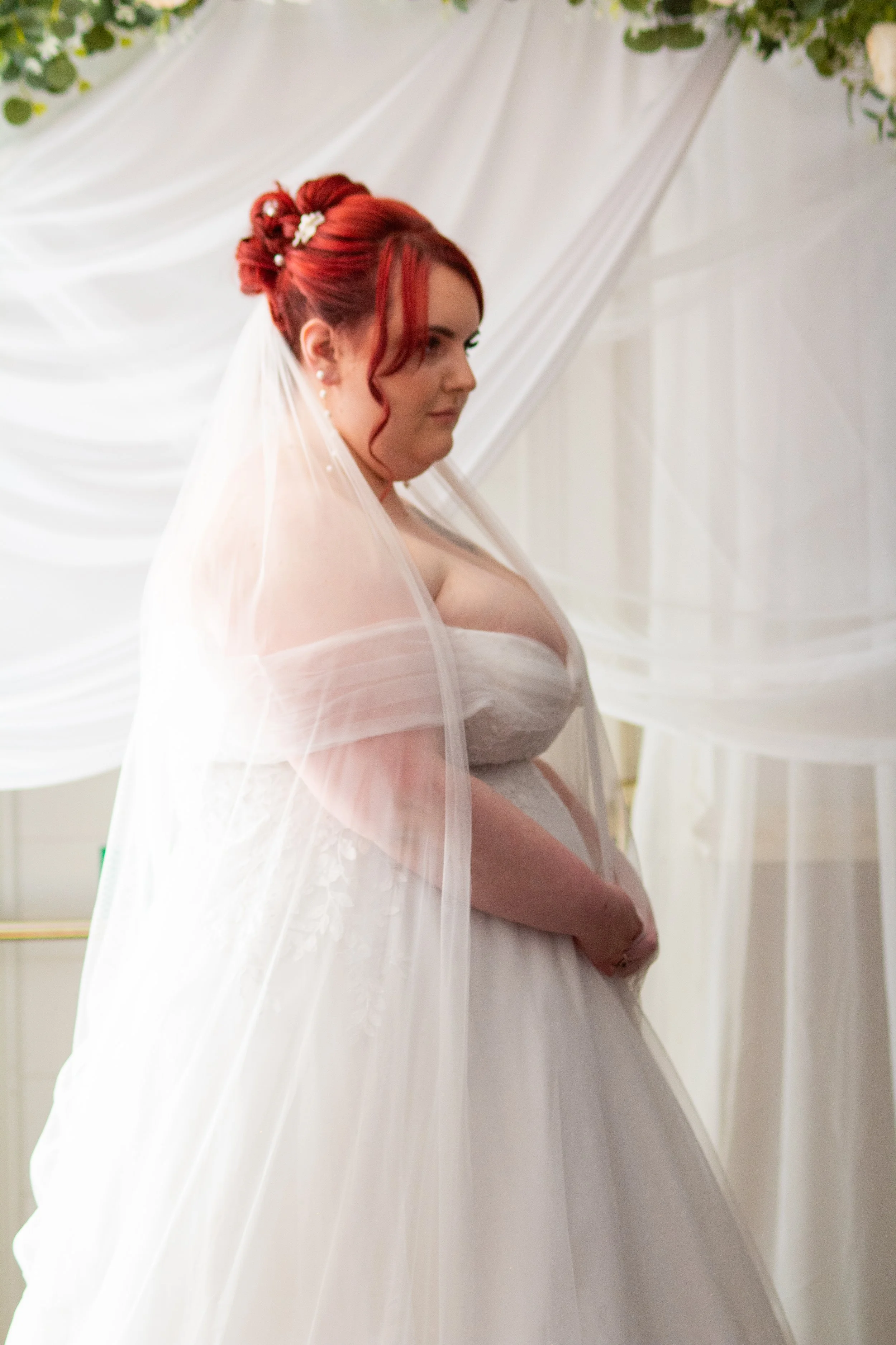 A bride with red hair styled in an updo, wearing a white wedding dress and veil, stands indoors against a backdrop of white draped fabric and greenery.