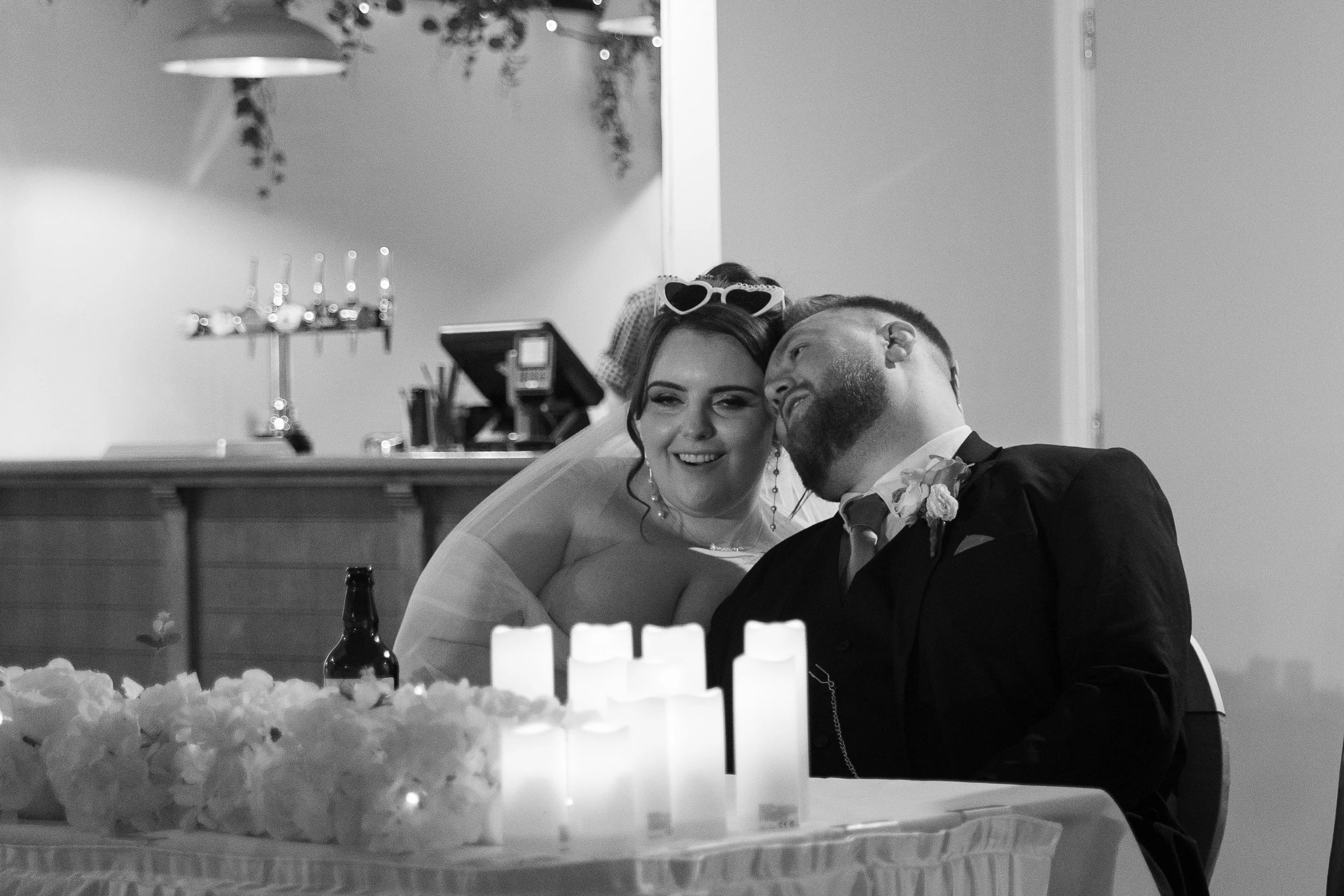 A bride and groom sitting at their wedding reception, smiling and leaning towards each other, with candles and flowers on the table in front of them.