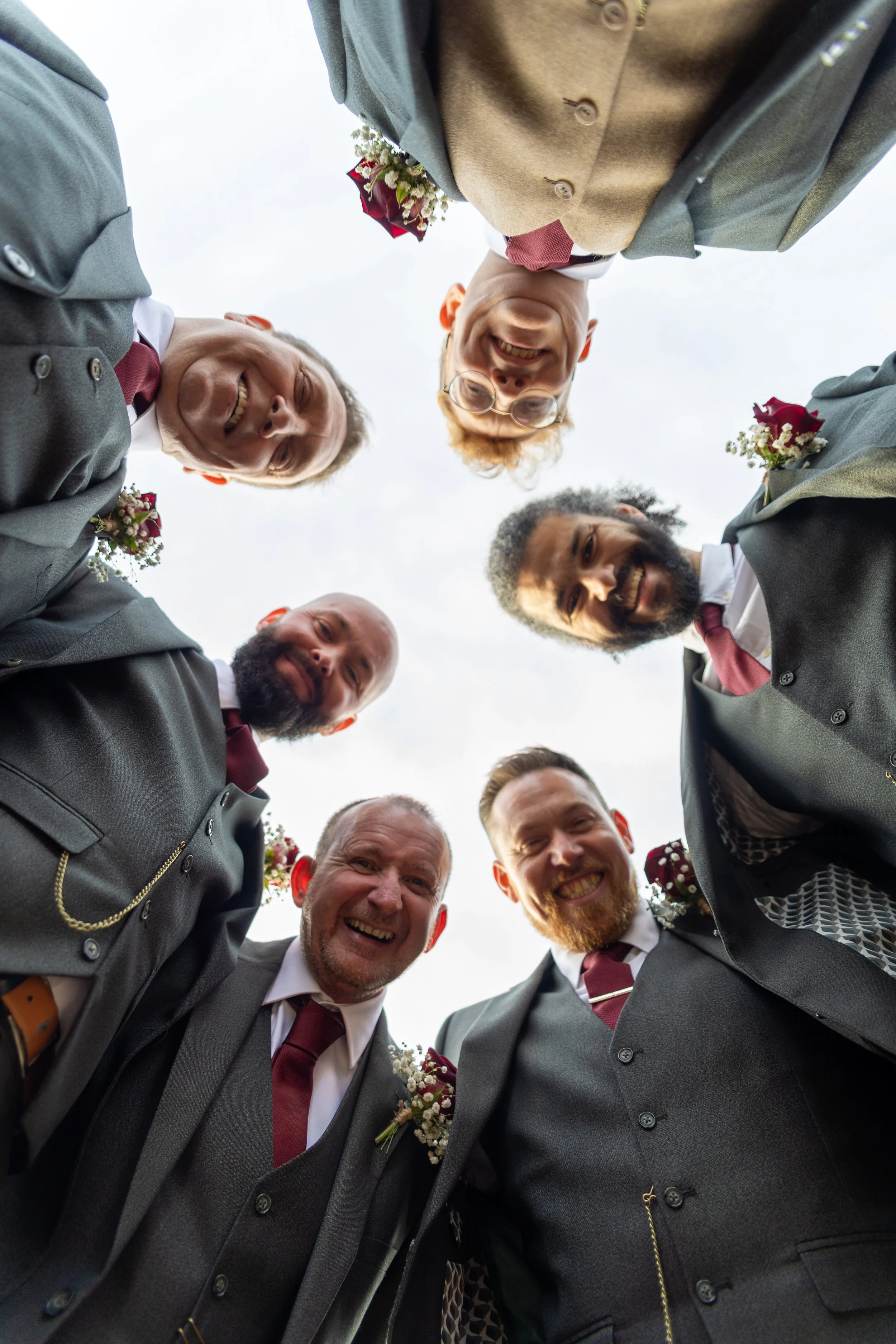 A group of people dressed in formal attire, looking down into the camera, forming a circle, smiling, with a cloudy sky in the background.