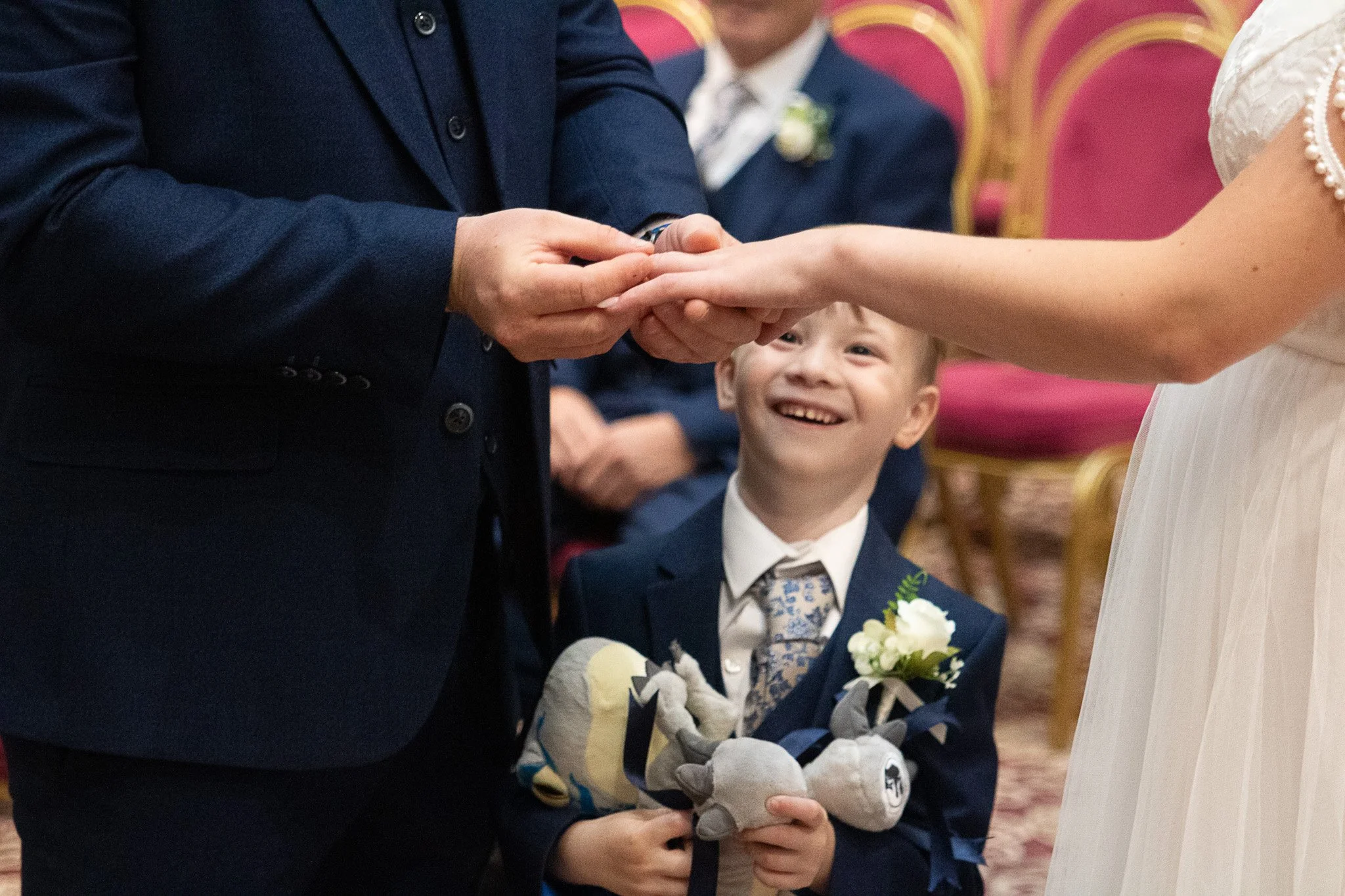 A young boy in a navy suit and tie is smiling and holding stuffed animals and a small bouquet of flowers, while watching a wedding ceremony where a groom and bride are exchanging rings.