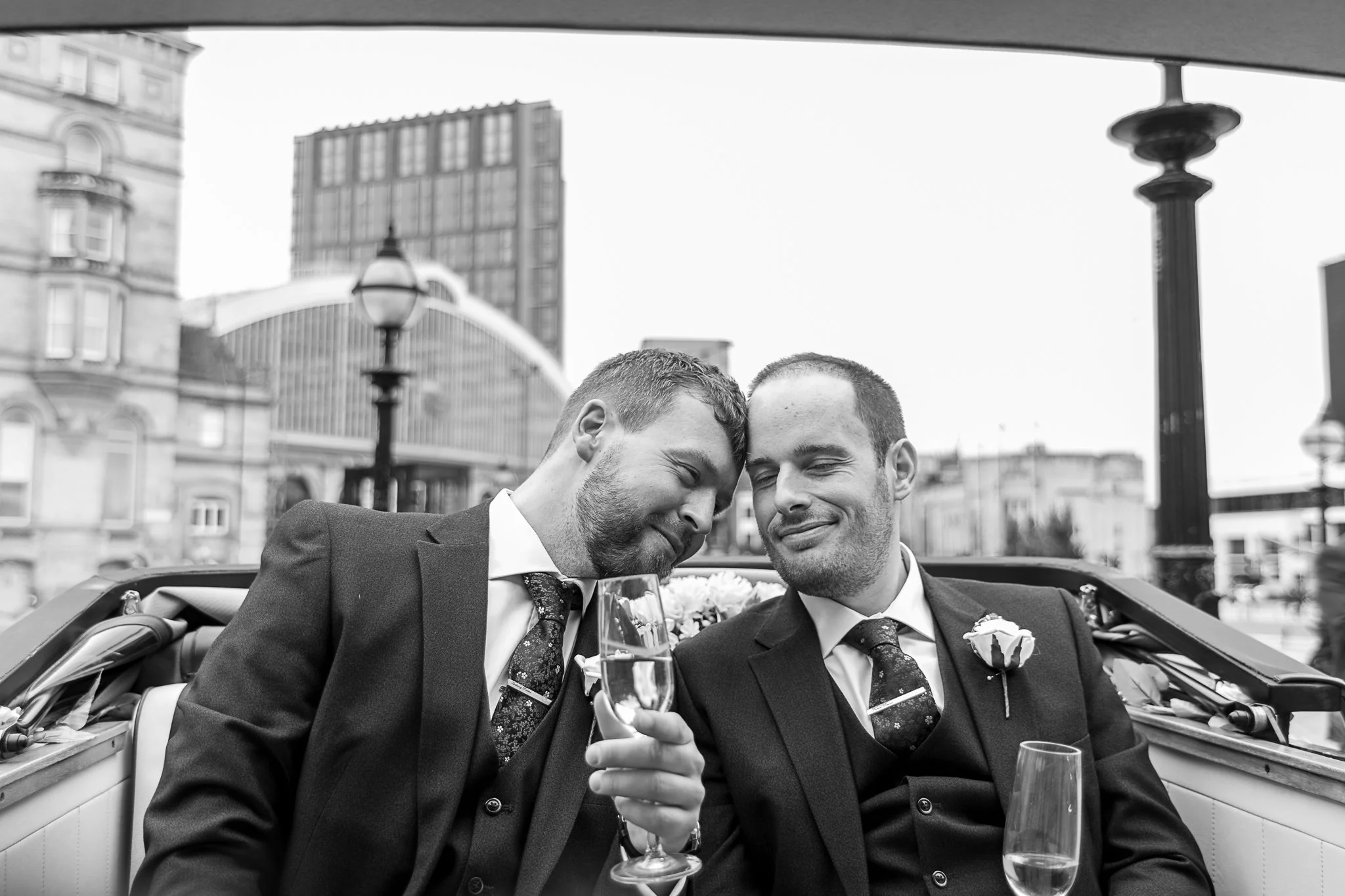 Two men in suits, sitting in a convertible car, sharing a close moment while holding champagne glasses, with city buildings and lampposts in the background.