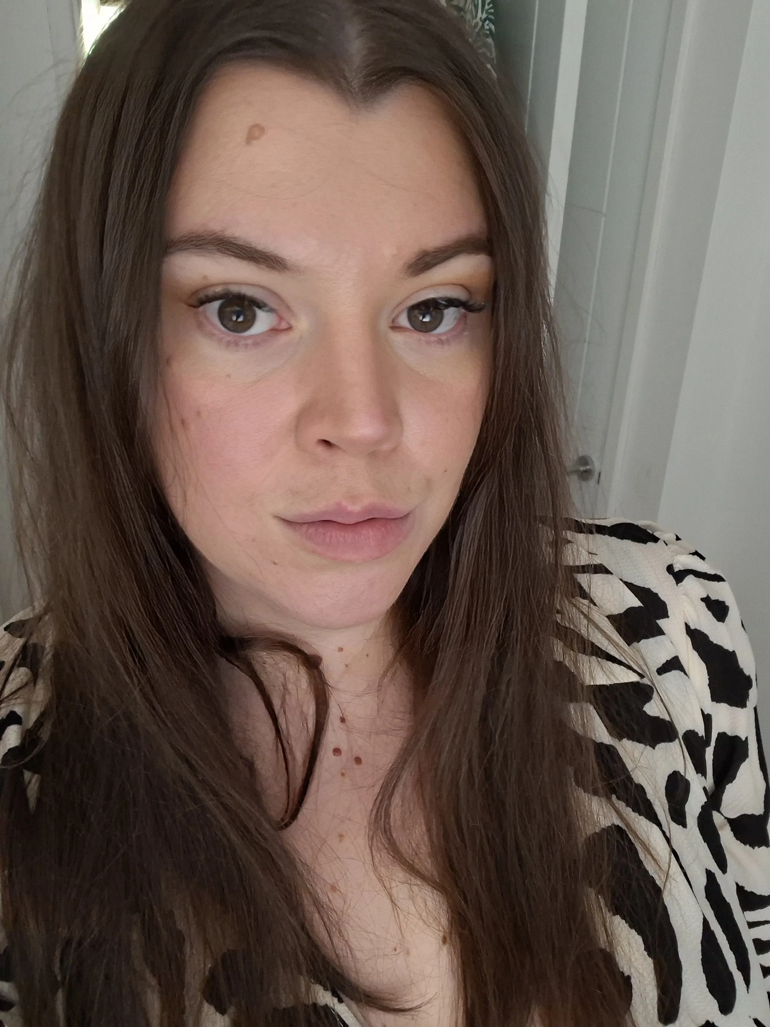 A woman with long brown hair and light makeup, wearing a black and white animal print top, standing indoors in front of a mirror.