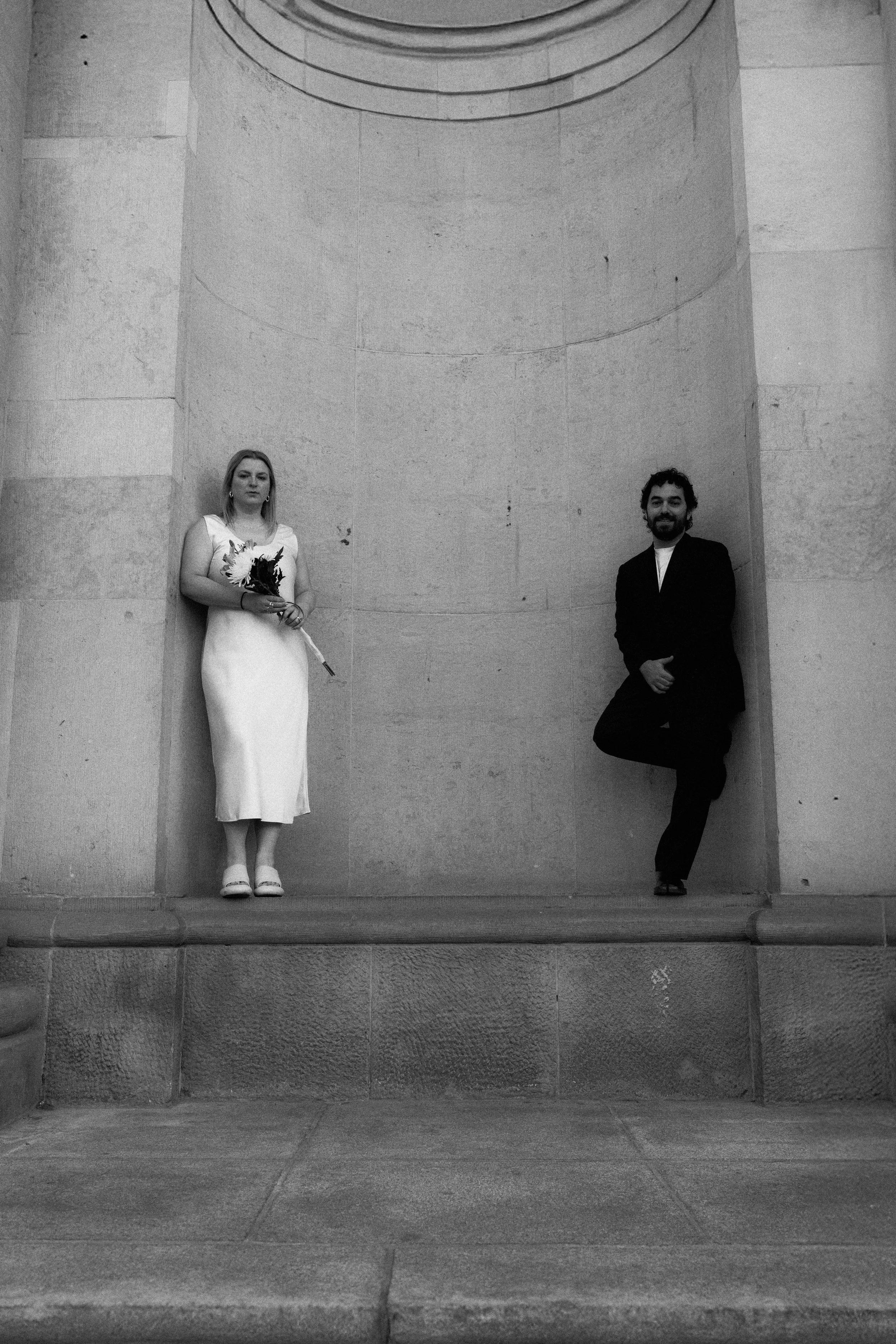 A black and white photo of a woman in a white dress holding a bouquet standing next to a man in a black suit with a beard, standing with one leg crossed and leaning against a large stone wall.