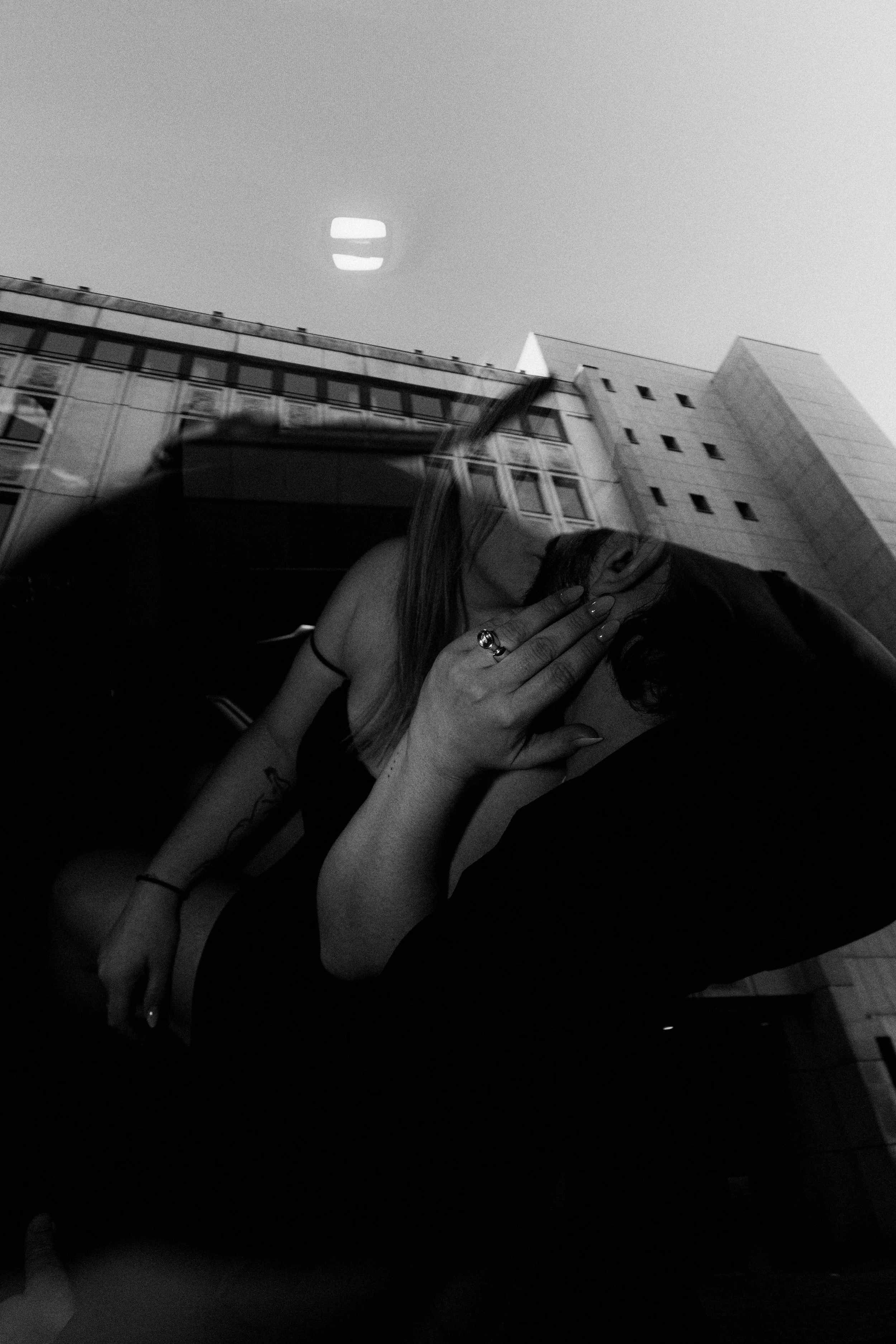 A black and white photo of a woman with long hair, wearing a sleeveless top, sitting inside a vehicle. She is covering her face with her hand, which has rings, and is shown through the reflection of a building with multiple windows on the glass windo