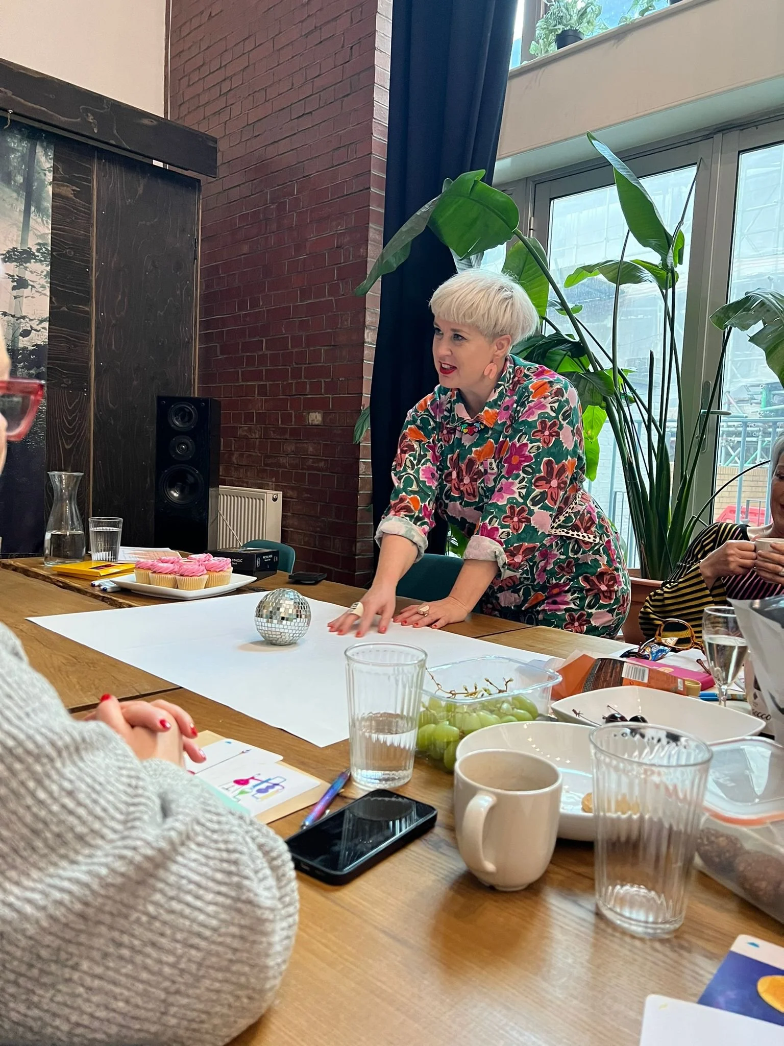 Woman with short blonde hair and floral shirt leaning on a table, speaking or presenting to a group, with a small disco ball and cupcakes on the table.