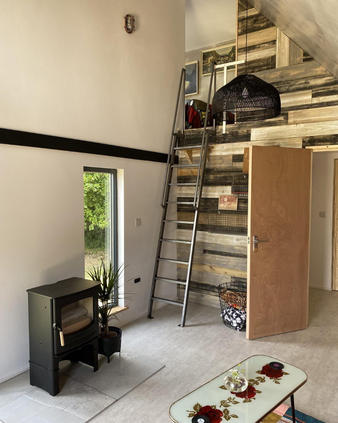 Interior of a modern living space with a small window, a potted plant, a black TV stand with a TV, a ladder leading to an upper level, and a wood-paneled wall behind the ladder. There is also a table with a glass top and floral design, and some artwork on the walls.