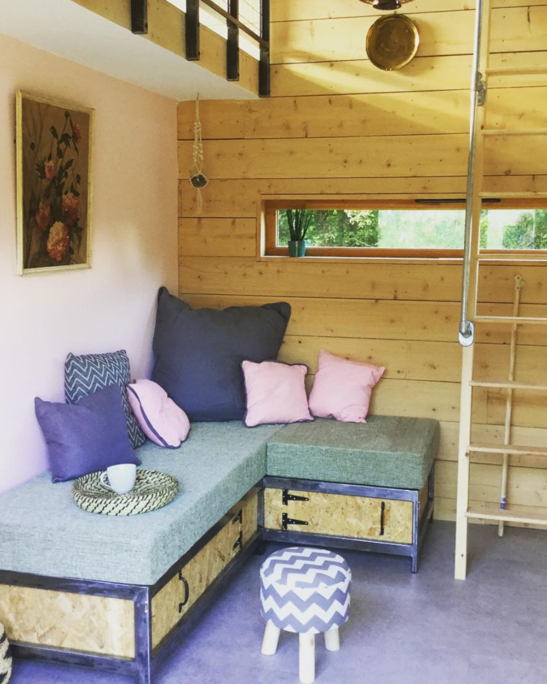 Cozy corner with a built-in sofa with cushions, a small stool with a zigzag pattern, a wooden ladder, a small horizontal window with a plant, and wall decor, in a room with wooden wall paneling.