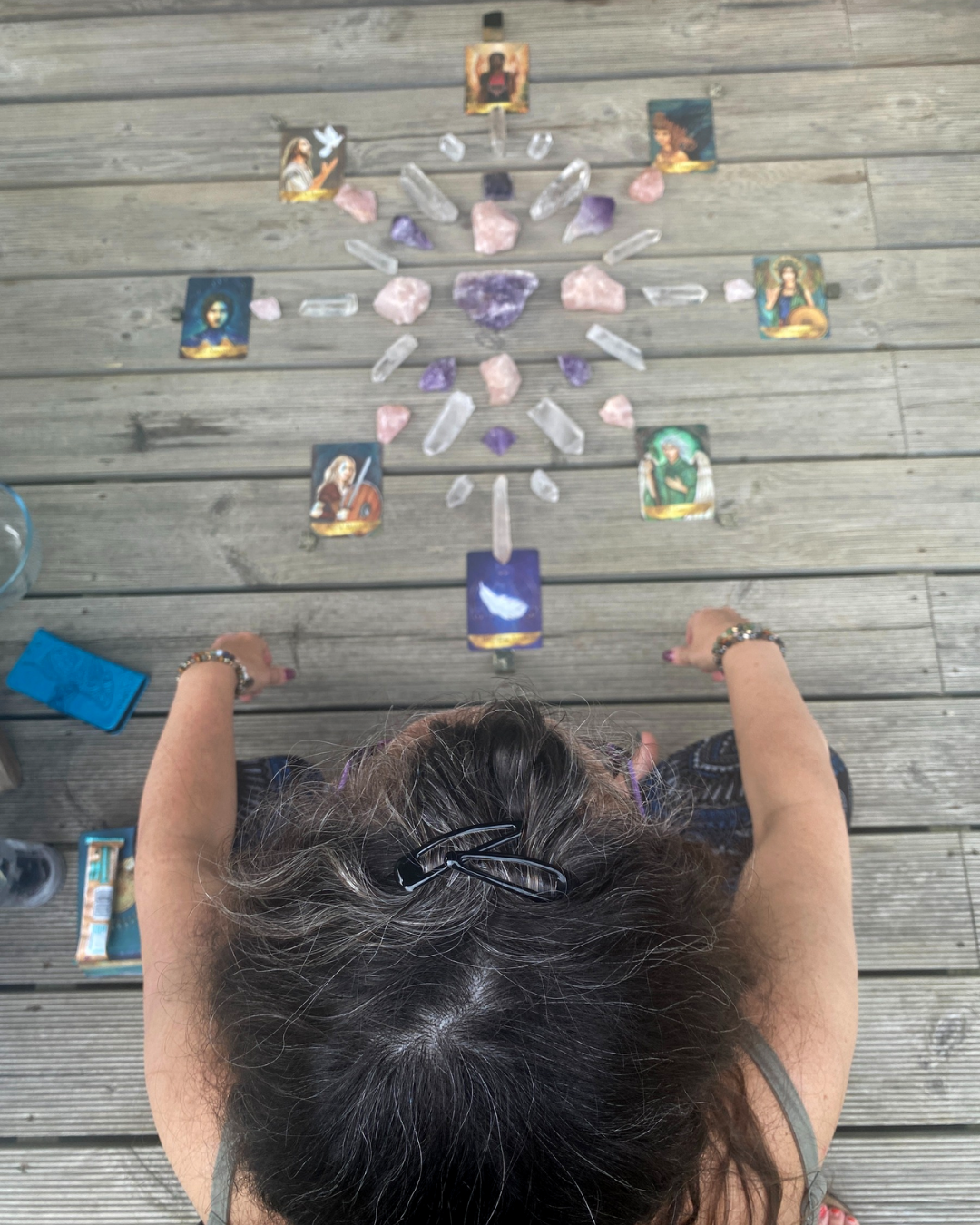 A woman with curly hair and a hair clip, viewed from above, is seated at a wooden table with various crystals and tarot cards arranged in a pattern. The tarot cards feature illustrated women and divine figures.