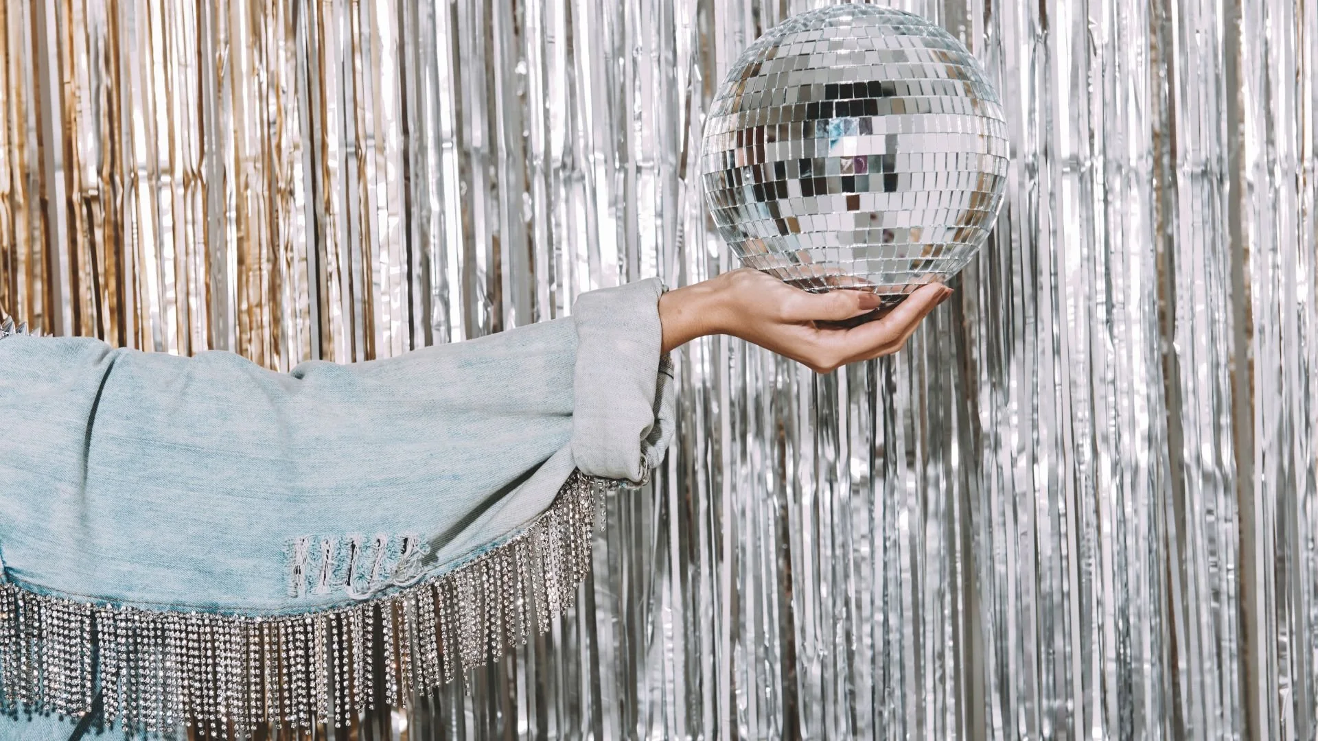 Person wearing a light denim jacket with fringe and rhinestones holding a disco ball in front of a silver tinsel backdrop.