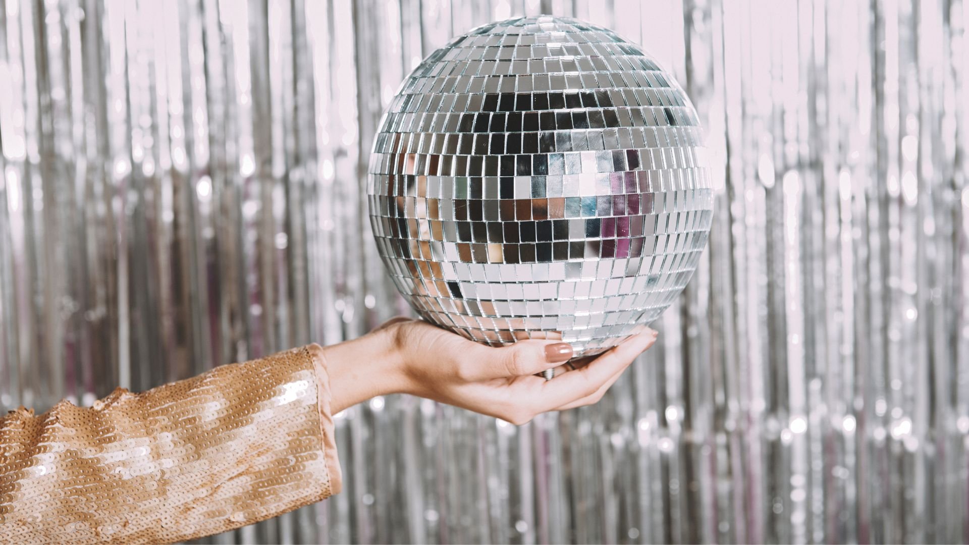 A person's hand holding a large disco ball in front of a metallic silver curtain.