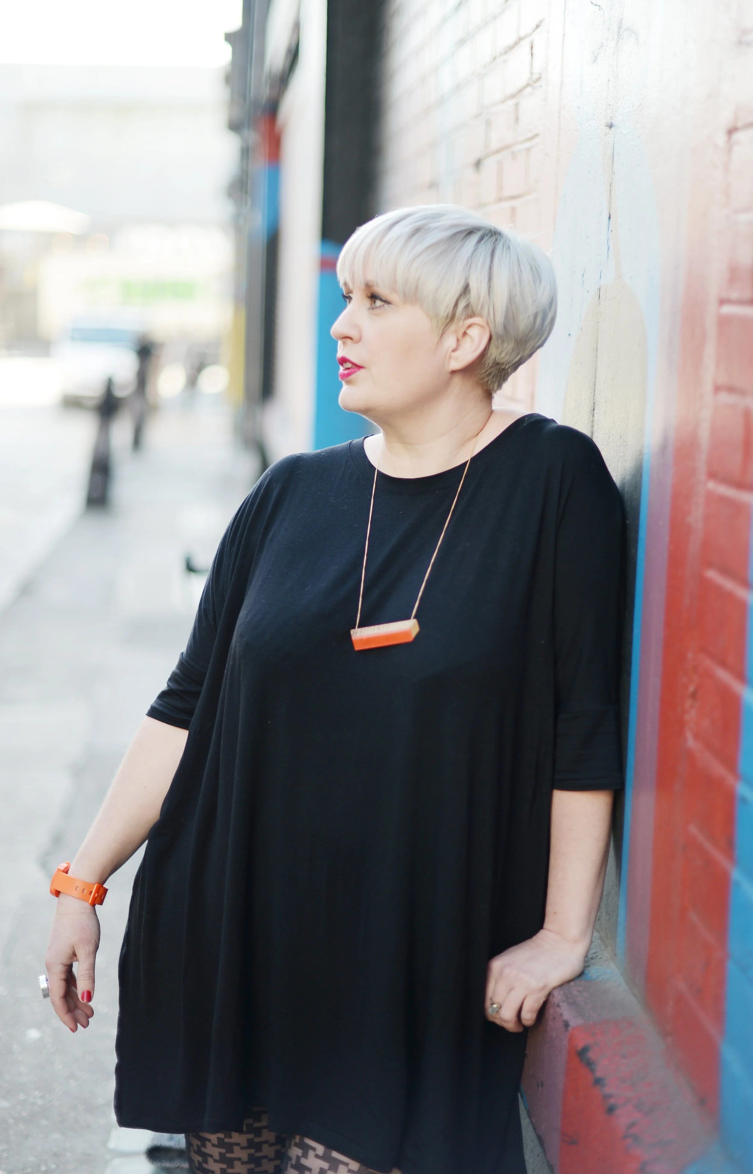 A woman with short blond hair in a black dress stands against a colorful brick wall on an urban street.