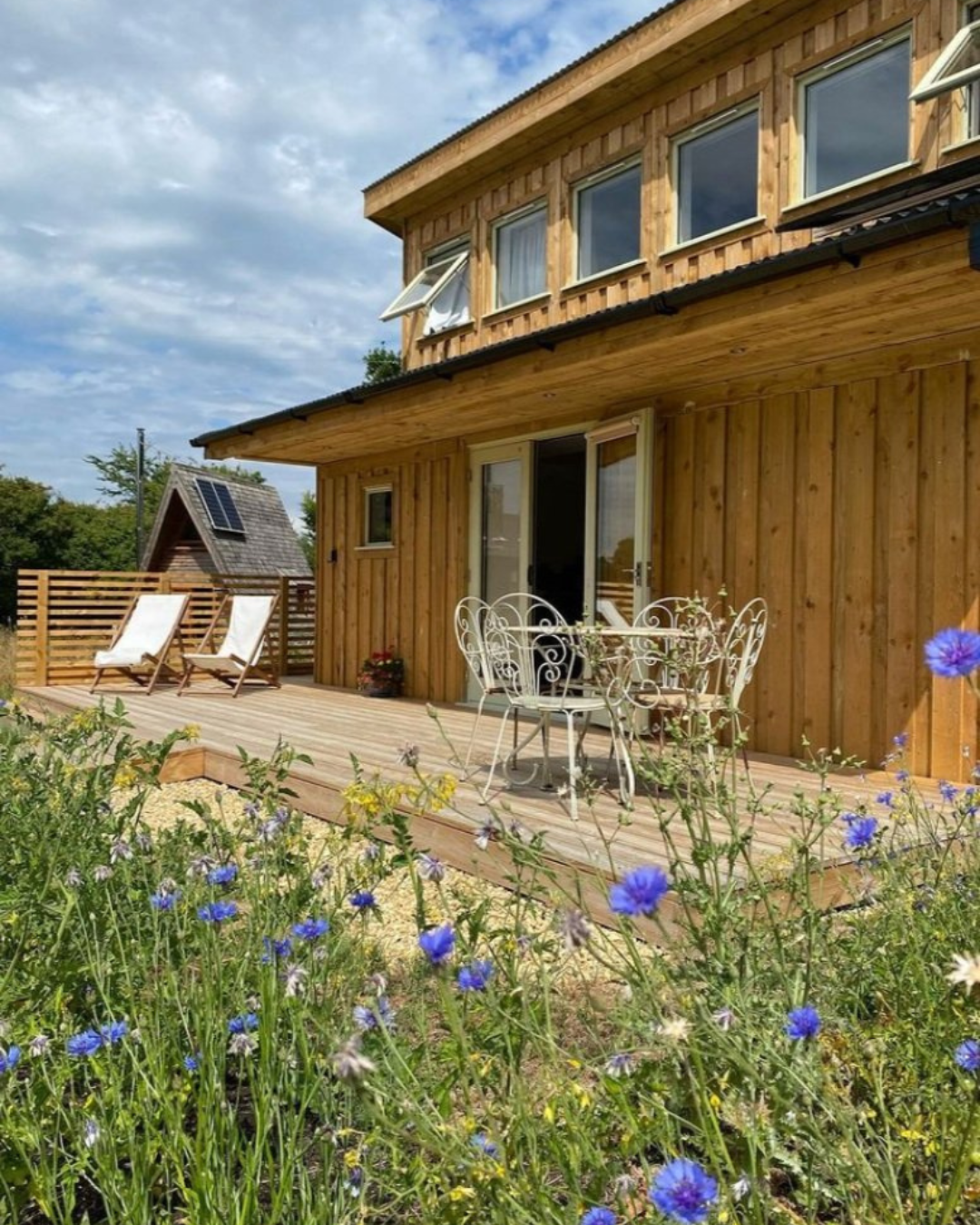 A wooden house with an outdoor deck, white wrought iron table and chairs, and lounge chairs surrounded by flowers under cloudy sky.