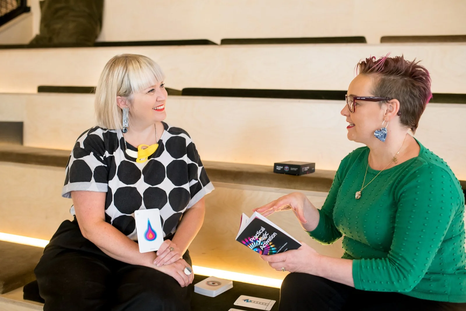 Two women having a conversation at an event, one holding a card with a colorful droplet graphic, and the other holding a booklet with the title 'Practical Application'.