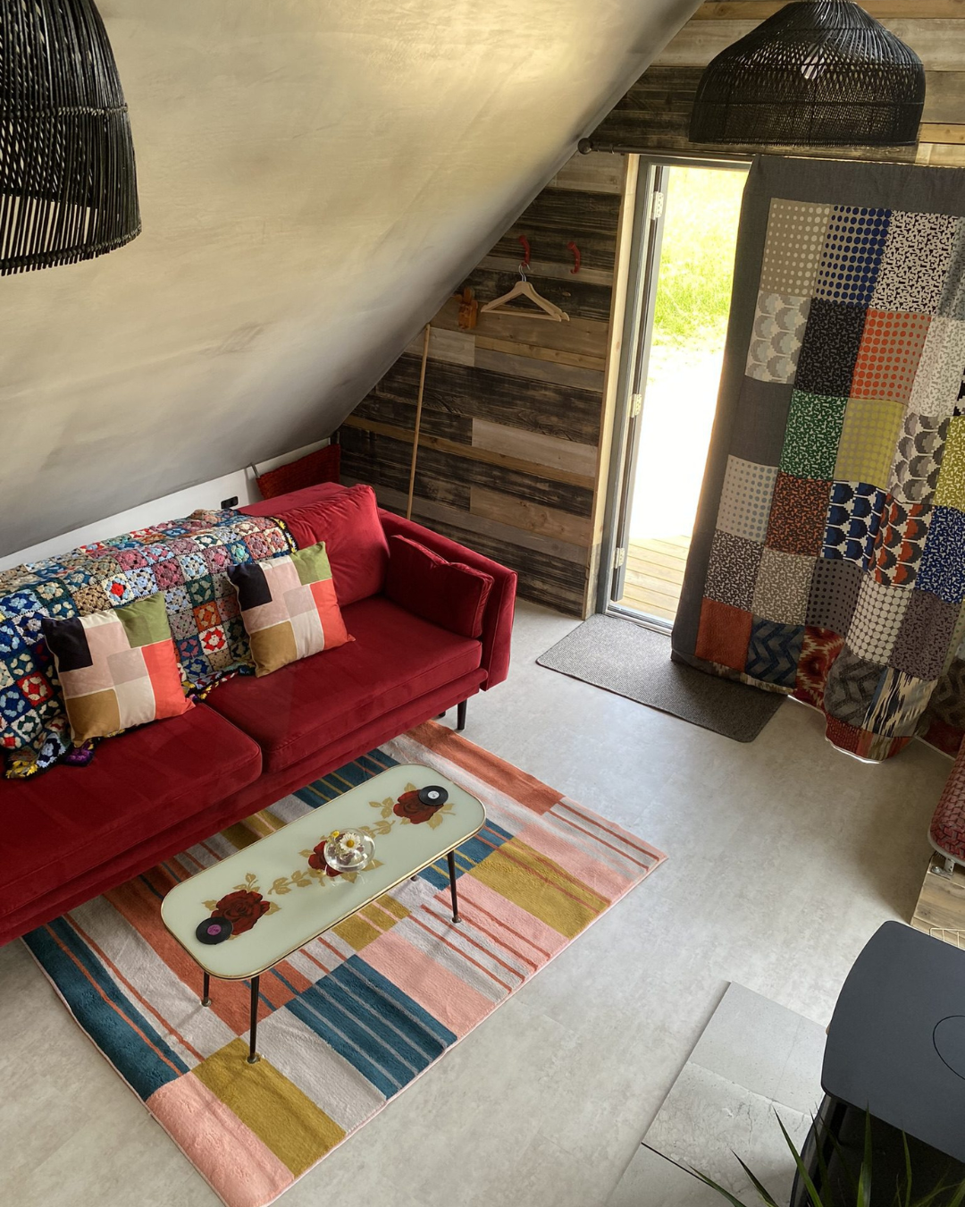 Cozy living room with a red velvet sofa, colorful patchwork pillows, a vintage floral coffee table, a multicolored geometric rug, and a patchwork curtain beside a small door opening to the outdoors. The walls are decorated with wooden planks, and black pendant lights hang from the ceiling.