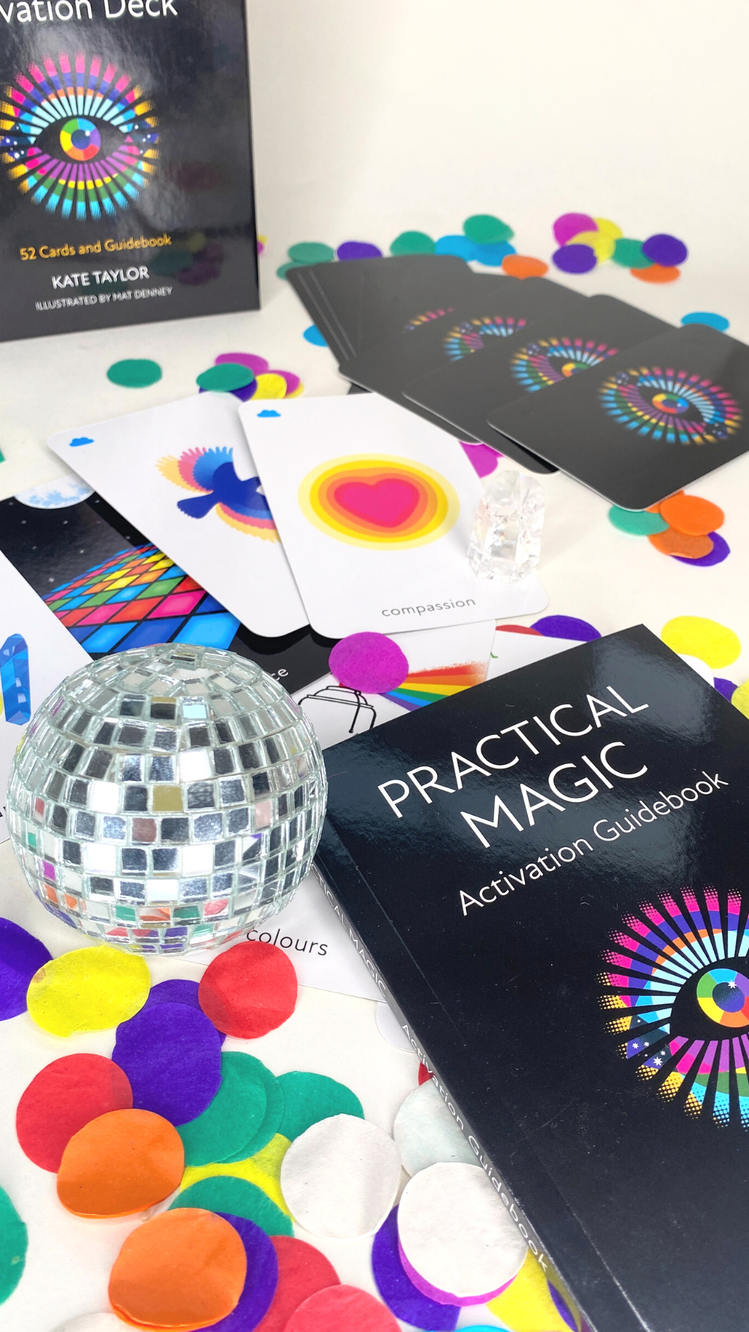 Colorful items on a table, including a black box labeled "Practical Magic Activation Guidebook," a deck of cards with a rainbow wheel design, a small disco ball, and colorful paper confetti.