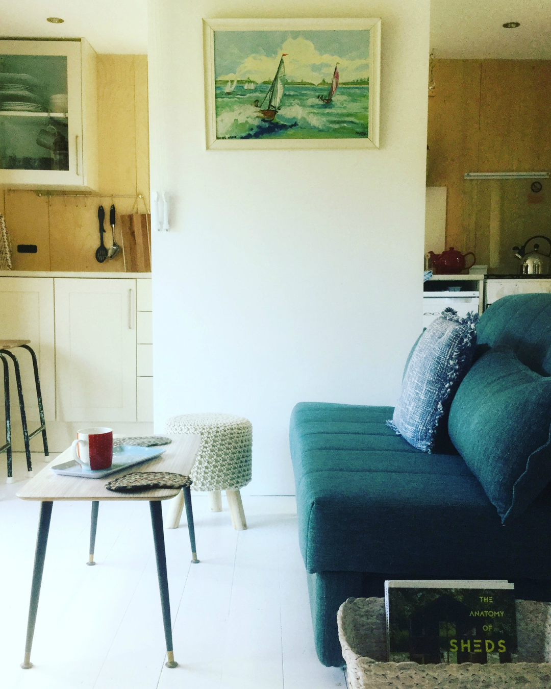 A cozy living room with a teal sofa, a small wooden coffee table with a red mug, a knitted pouf, a framed painting of sailboats on the water, and a partial view of a kitchen in the background.