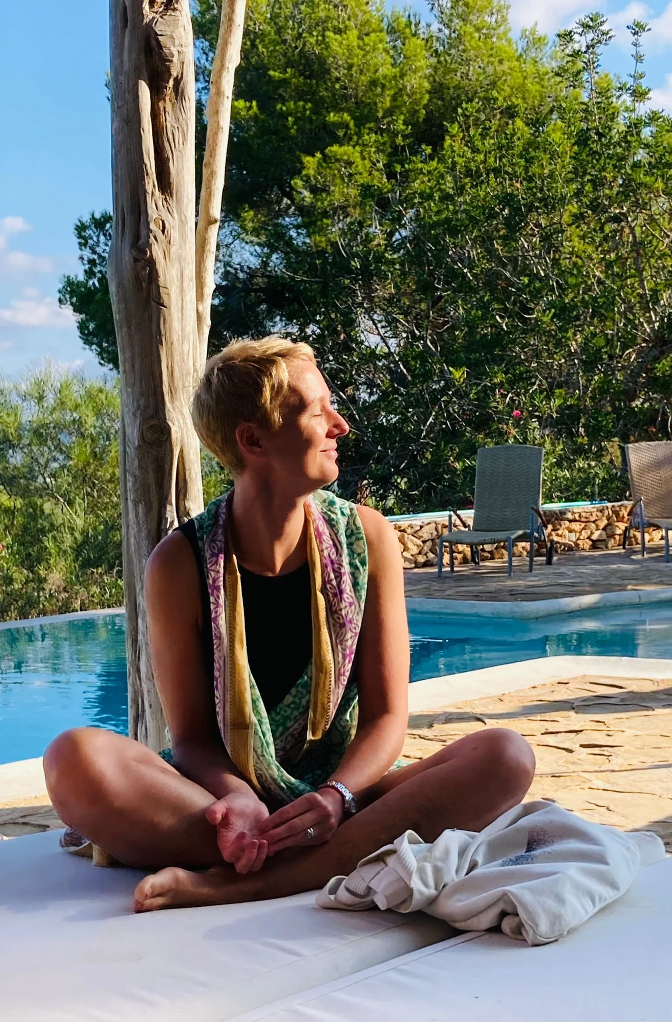 A woman with short blonde hair sitting cross-legged outdoors near a swimming pool, smiling and enjoying the sunlight, with trees and outdoor chairs in the background.