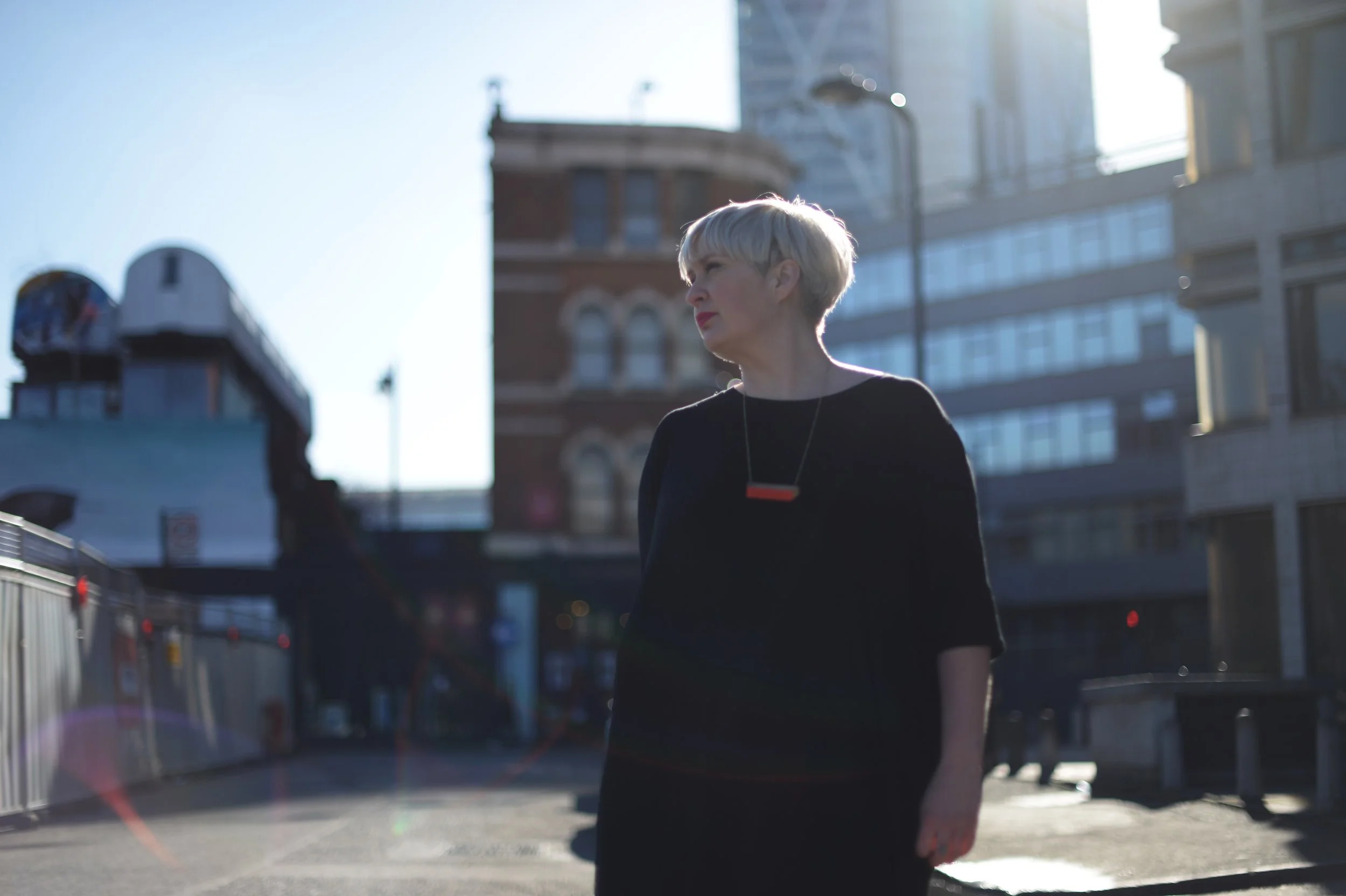 A woman with short blonde hair standing on a city street during daytime, wearing a black top and a red and black necklace, with modern buildings in the background and sunlight creating a silhouette effect.