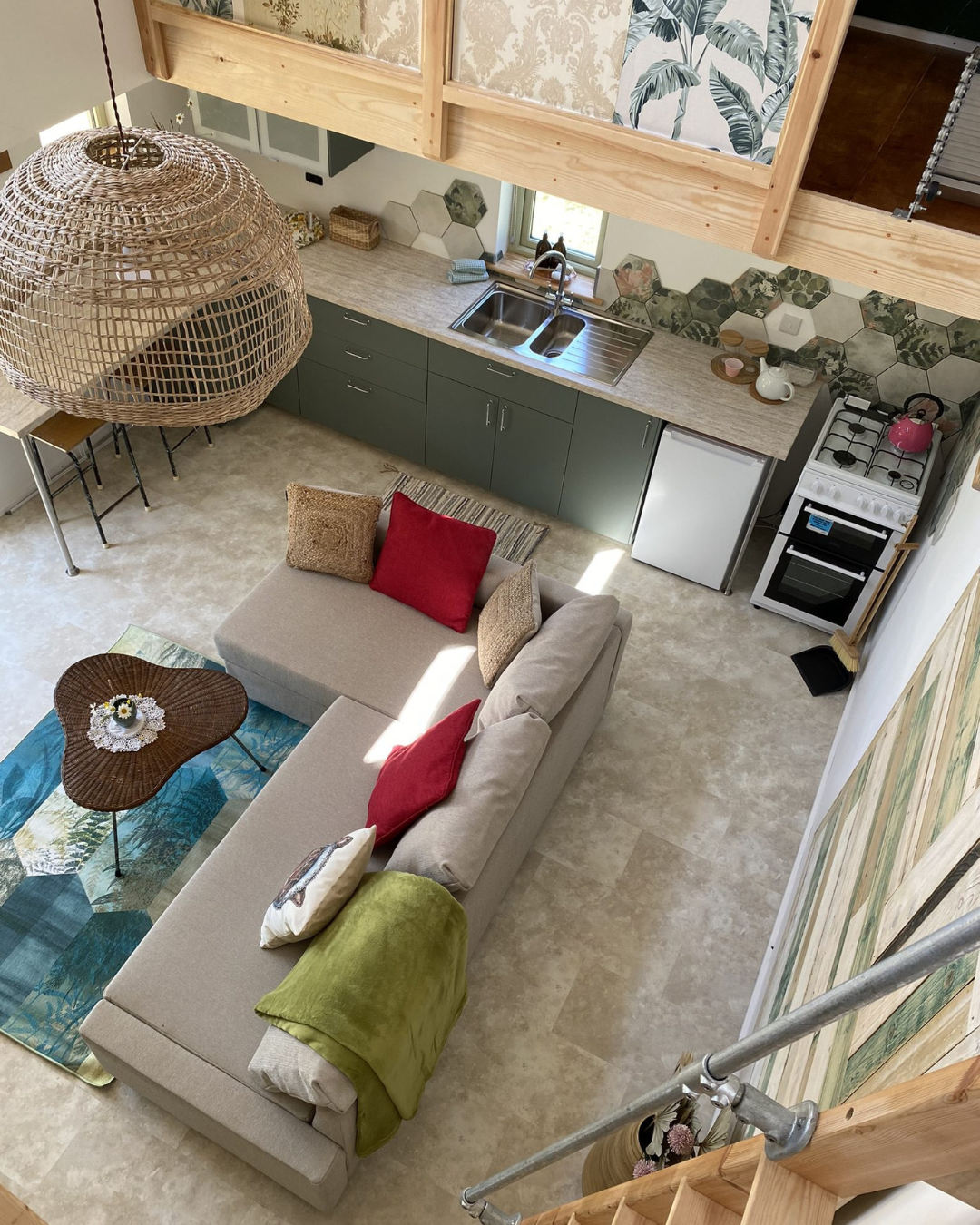 A cozy open-concept living space with a beige sectional sofa adorned with red, beige, and green pillows, a small wooden coffee table with a flower arrangement, and a colorful rug. The kitchen area features green cabinets, a beige countertop, a stainless steel sink beneath a window, and a white stove with a pink kettle. Hexagonal tiles with botanical patterns create a backsplash behind the sink. A wooden balcony railing partially covers the view from above, with a woven hanging light fixture overhead.