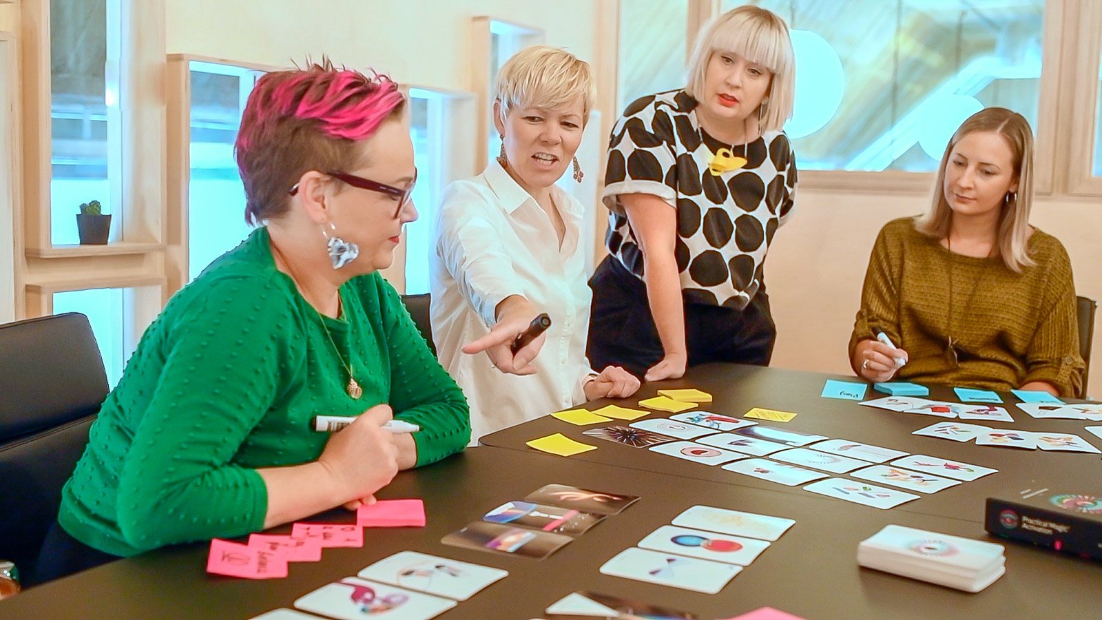 Four women playing a card game around a table, with colorful cards and sticky notes, inside a well-lit room with wooden accents.