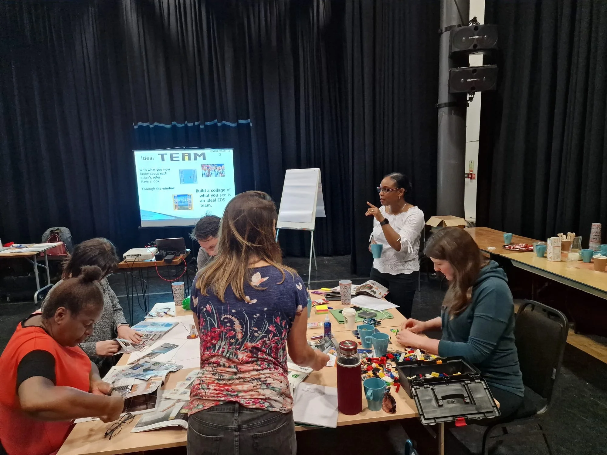 A group of people gathered around a table in a workshop or training session with a woman standing and speaking, gesturing with her right hand while holding a cup in her left. The table is cluttered with papers, cups, and a LEGO set. In the background, there's a screen displaying a presentation slide titled 'TEAM' and various chairs and tables.
