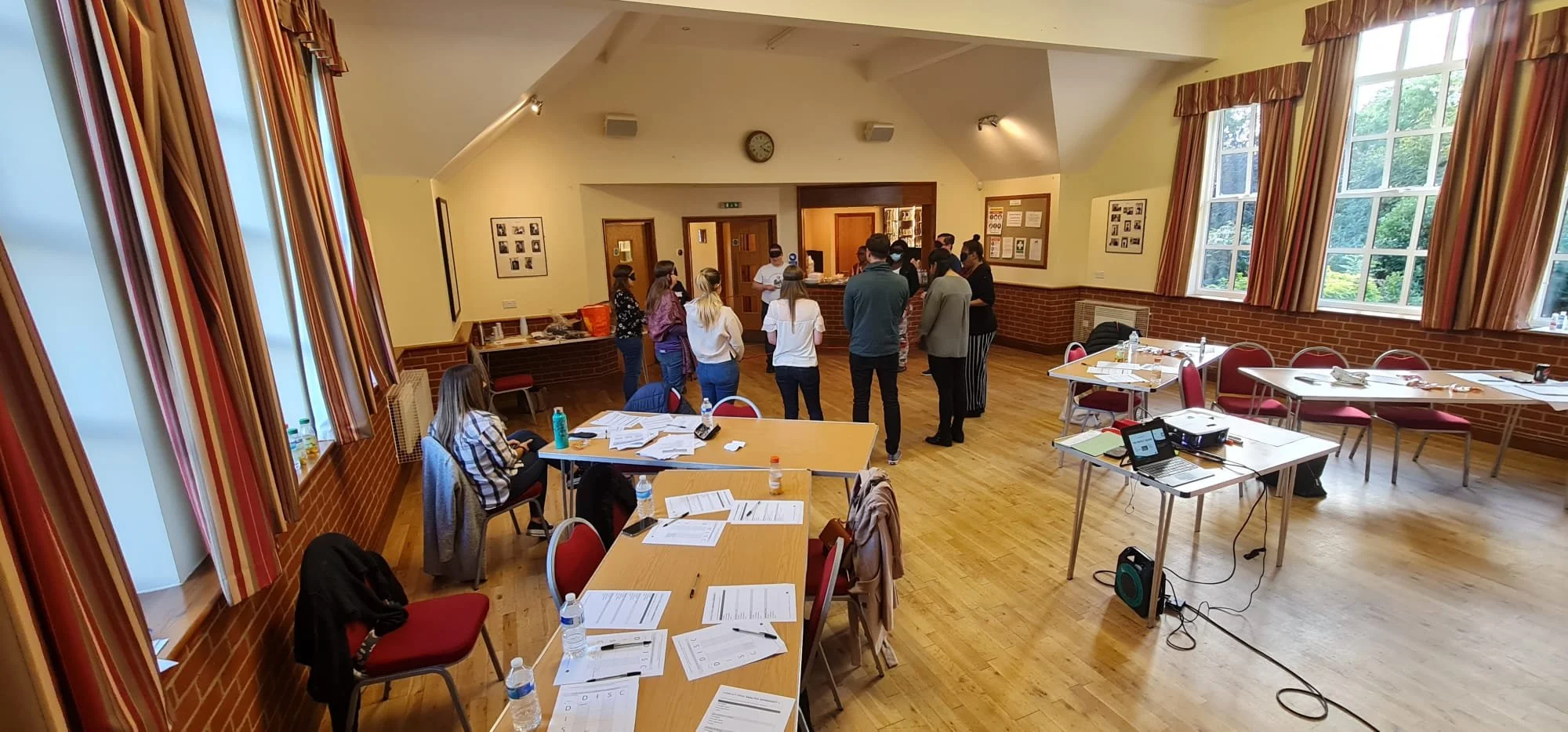 A group of people standing in line at a reception desk inside a well-lit room with large windows, wooden floors, and red chairs around tables covered with papers, water bottles, and other materials.
