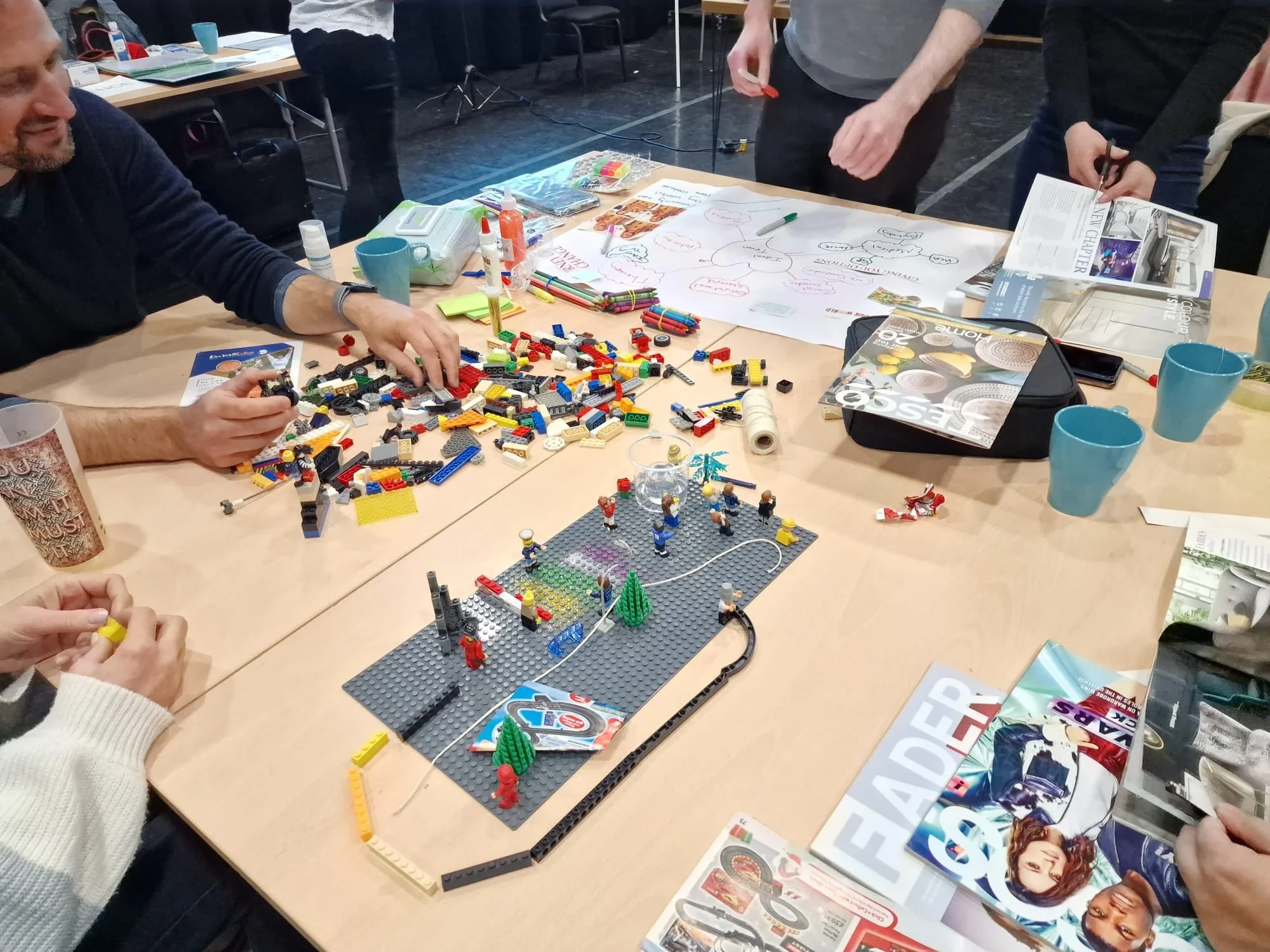 People working on a LEGO project at a table with various LEGO pieces, magazines, and papers, in a meeting or workshop setting.