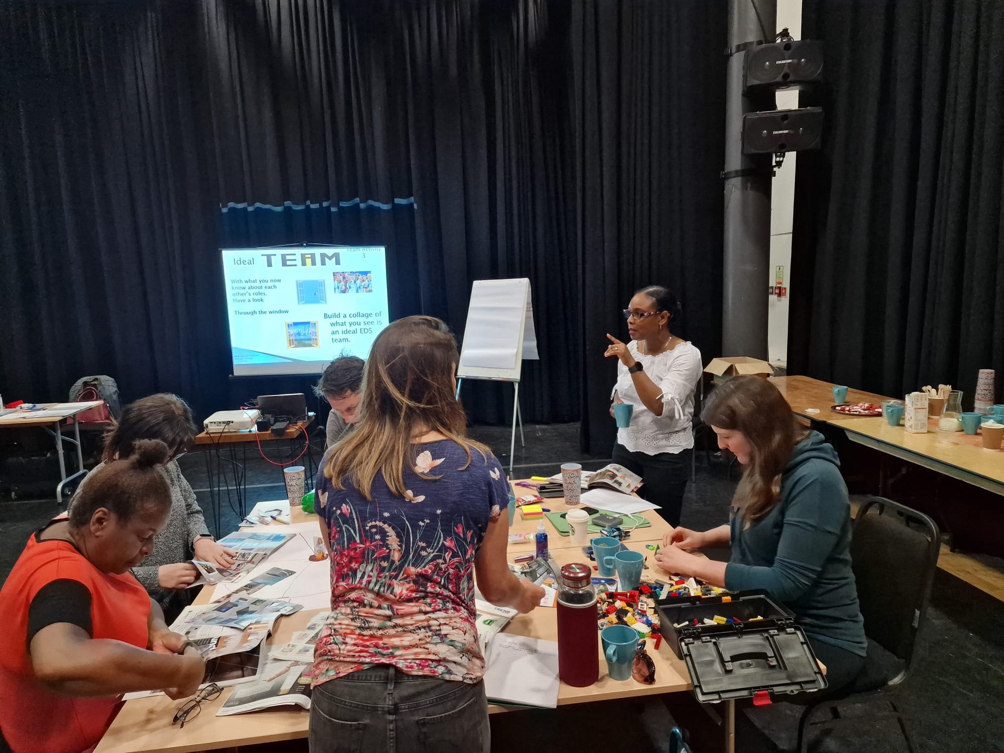 A group of women around a table in a workshop setting, with a presenter standing and pointing at a screen that displays a presentation about team building, surrounded by cups, papers, and various workshop materials in a black-curtained room.