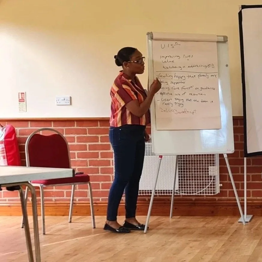 A woman wearing glasses, a striped shirt, and dark pants standing next to a whiteboard with handwritten notes in a room with a brick half-wall and wooden floors. She appears to be giving a presentation or lecture.