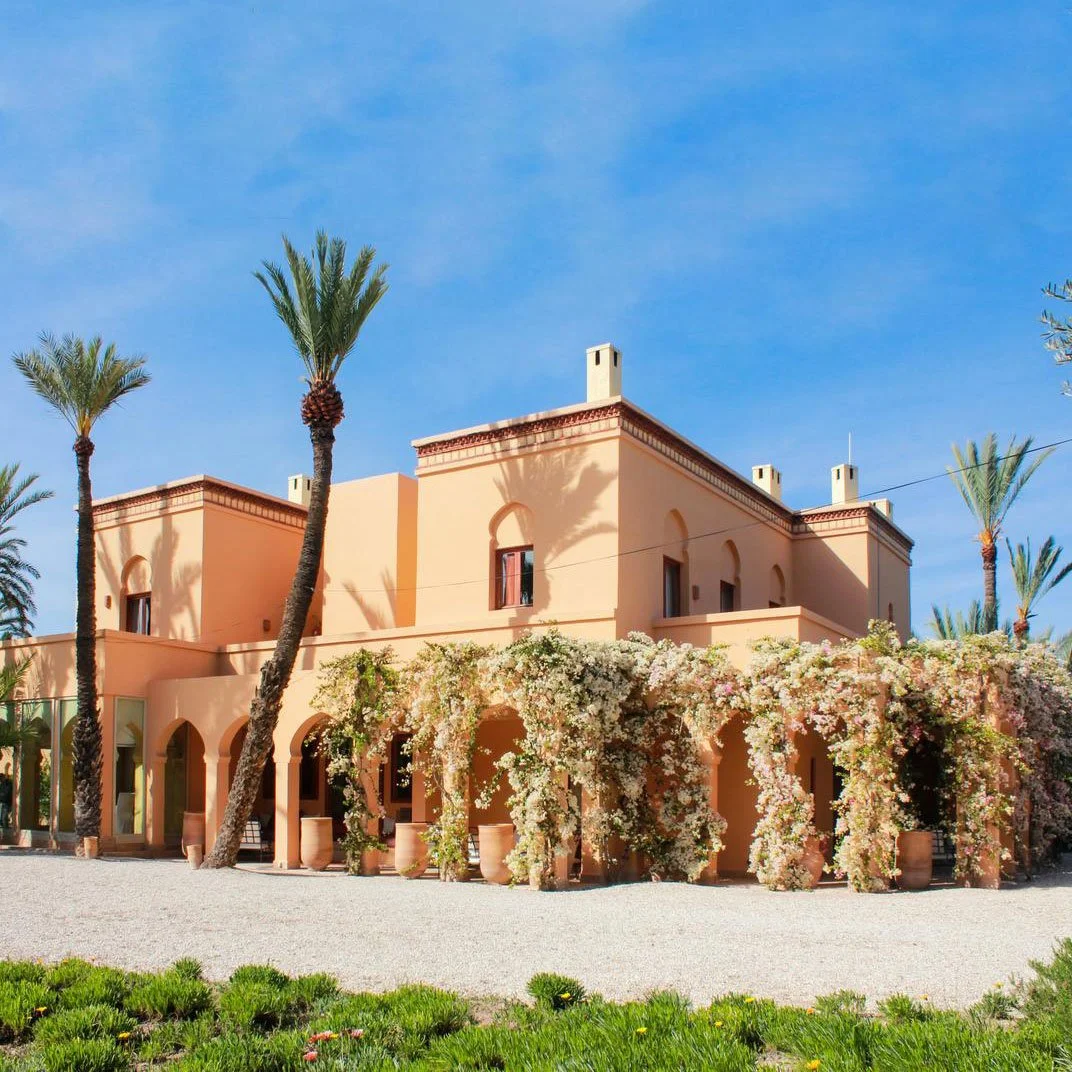 A peach-colored villa with lush flowering vines, palm trees, and a gravel courtyard under a blue sky.