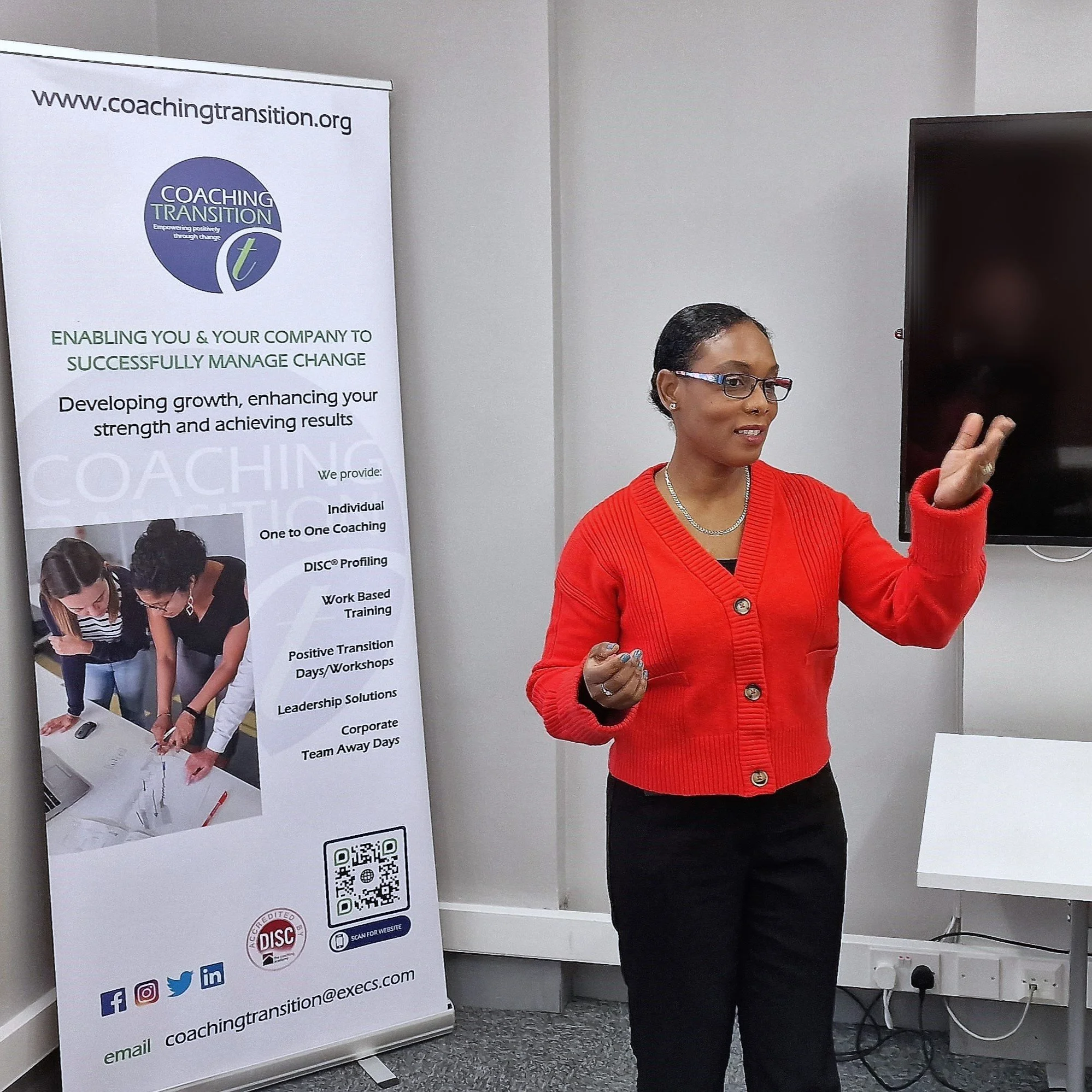 A woman in a red sweater and black pants giving a presentation in a conference room next to a banner for coaching transition services.