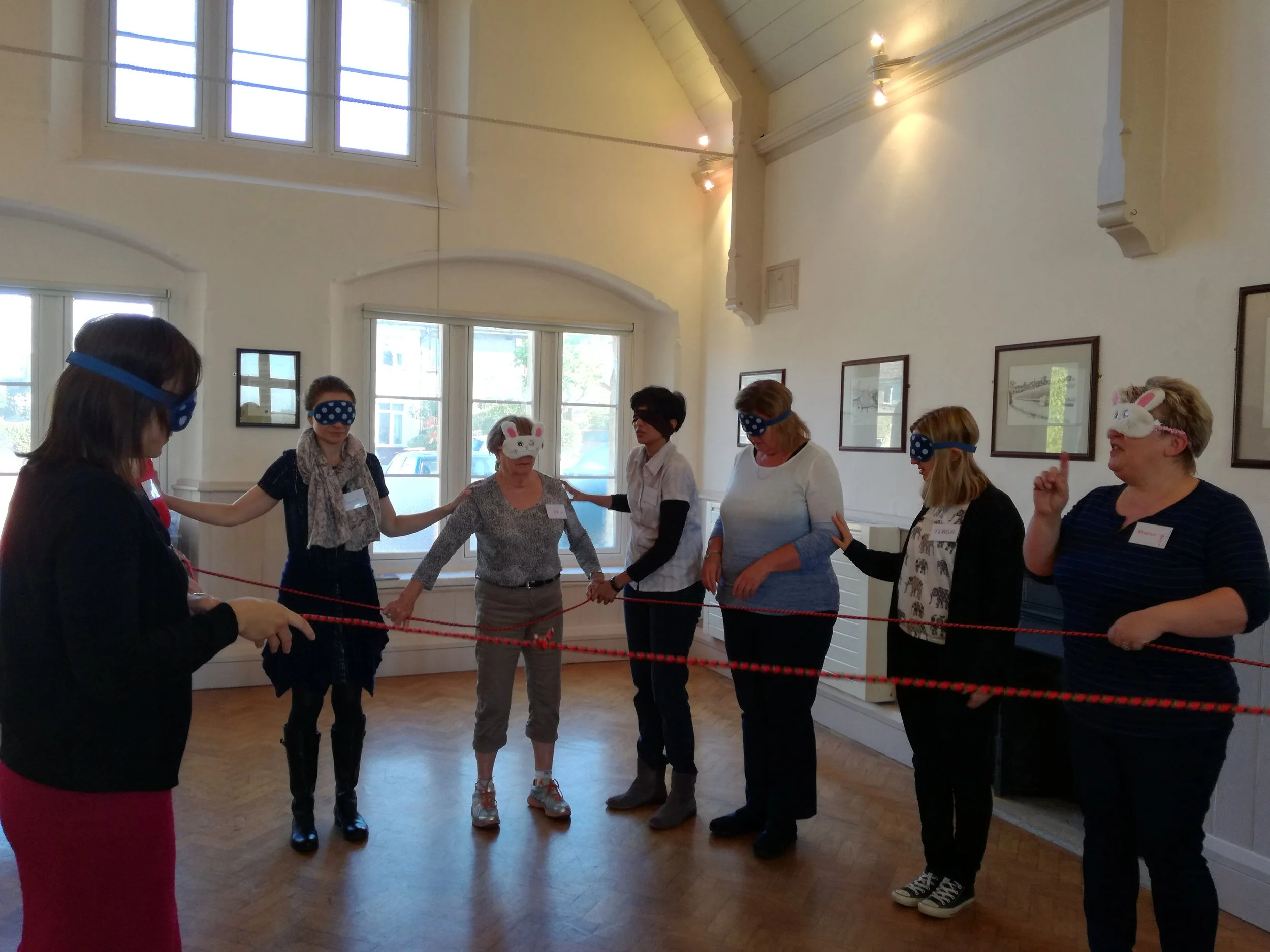 Group of people participating in a team-building activity, wearing blindfolds and masks, holding a red rope together in a room with framed pictures on the wall and large windows.