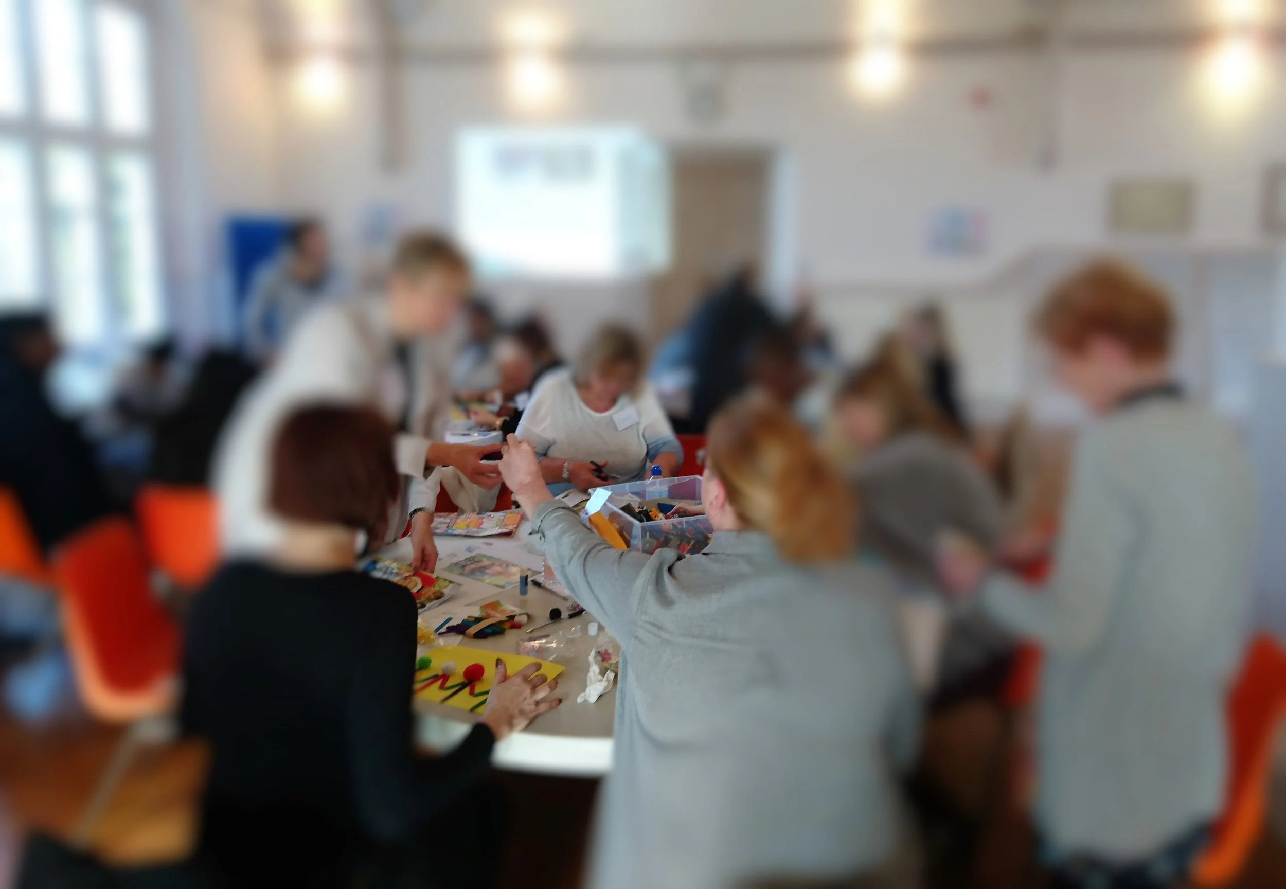 People gathered around a table participating in a group activity or craft in a brightly lit room, possibly a classroom or workshop setting.