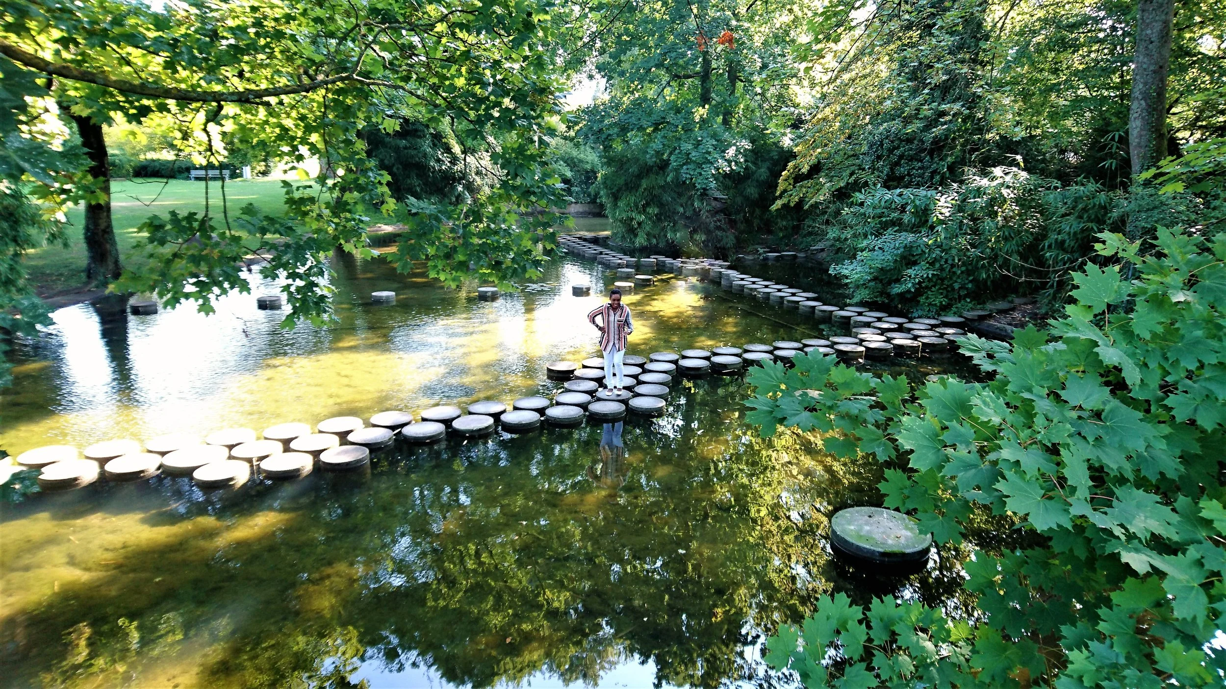 A person standing on a circular stepping stone pathway across a shallow pond in a lush green park, with overhanging trees and reflections on the water.