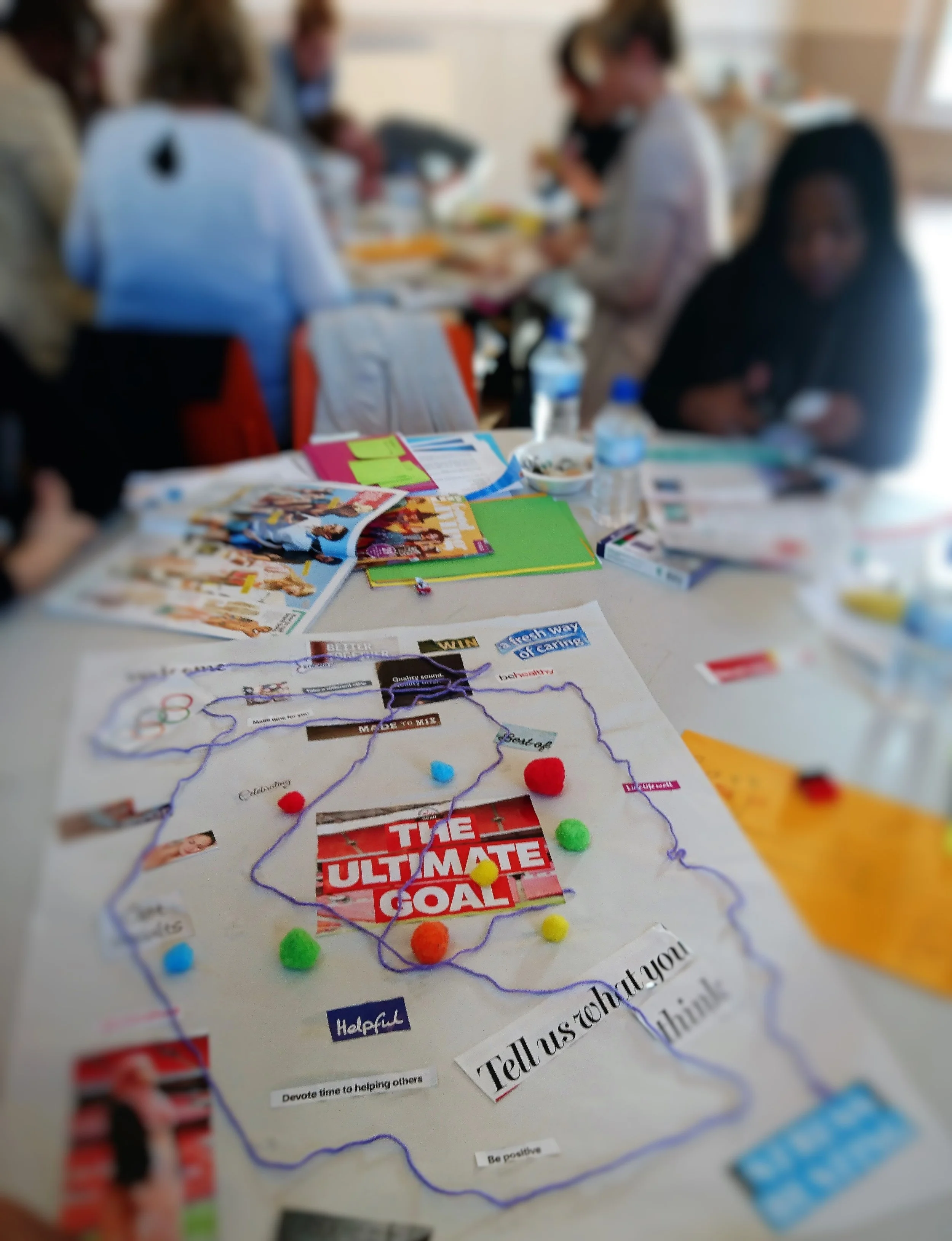Group of people gathered around a table, working on a project with colorful papers, magazines, and craft supplies, with a poster featuring a game or activity in the foreground.