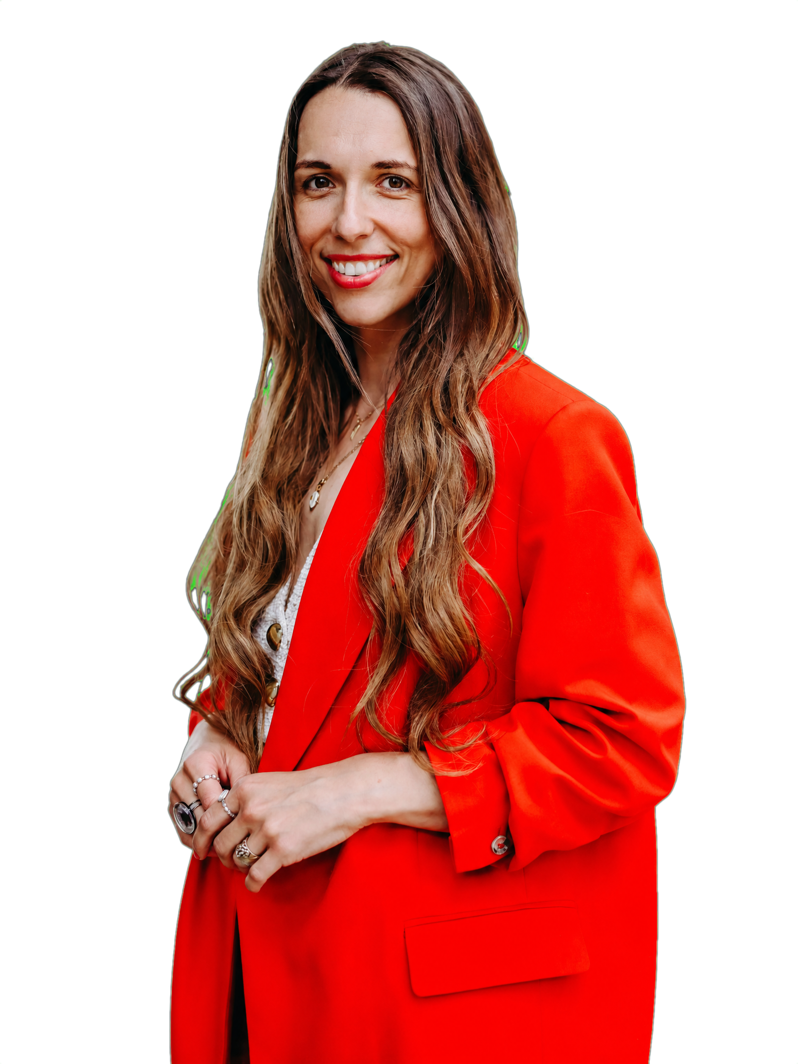 A woman with long brown wavy hair, smiling, wearing red lipstick, a red blazer, and several rings, standing with her arms crossed.